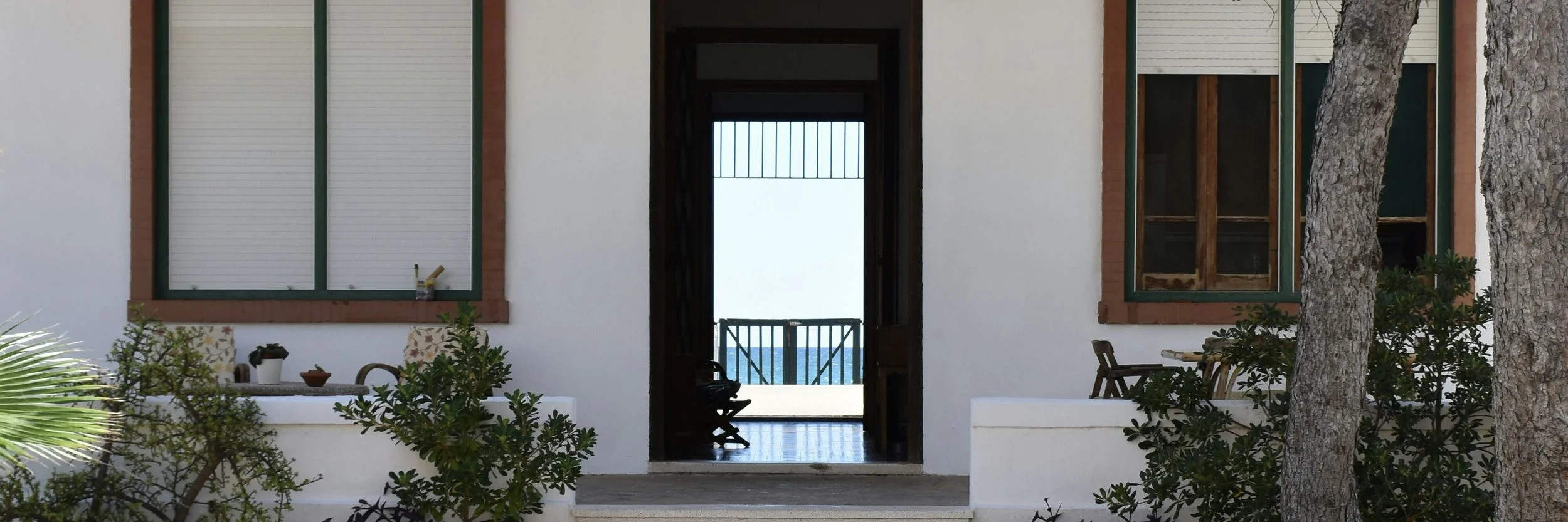 Front view of a house with open door leading to a balcony, ocean view, two large windows with green frames, outdoor plants, and trees.