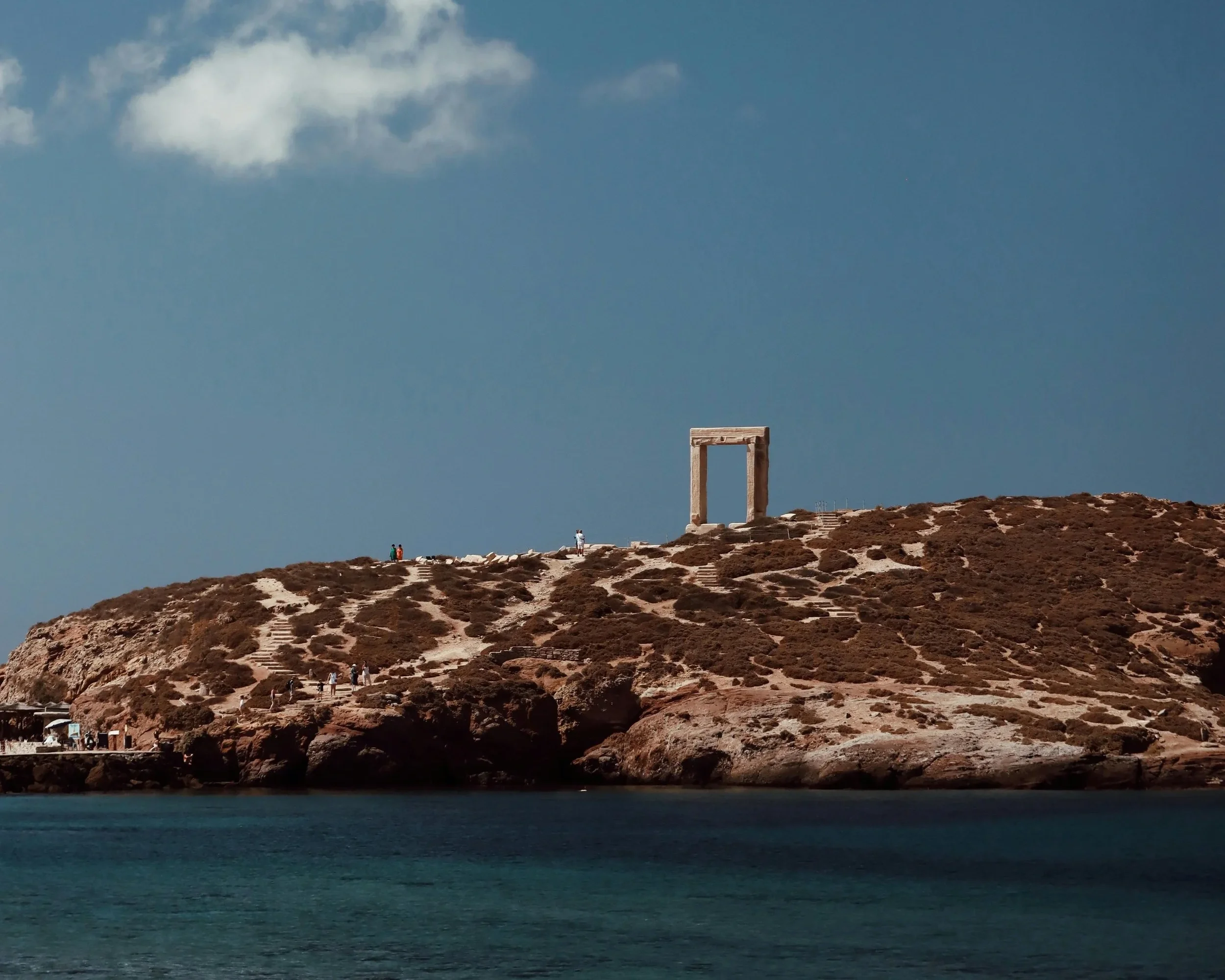 A rocky hillside near the ocean with a large, rectangular, stone arch structure on top, and a few people walking along a staircase and trail on the hill. The sky is mostly clear with some clouds.
