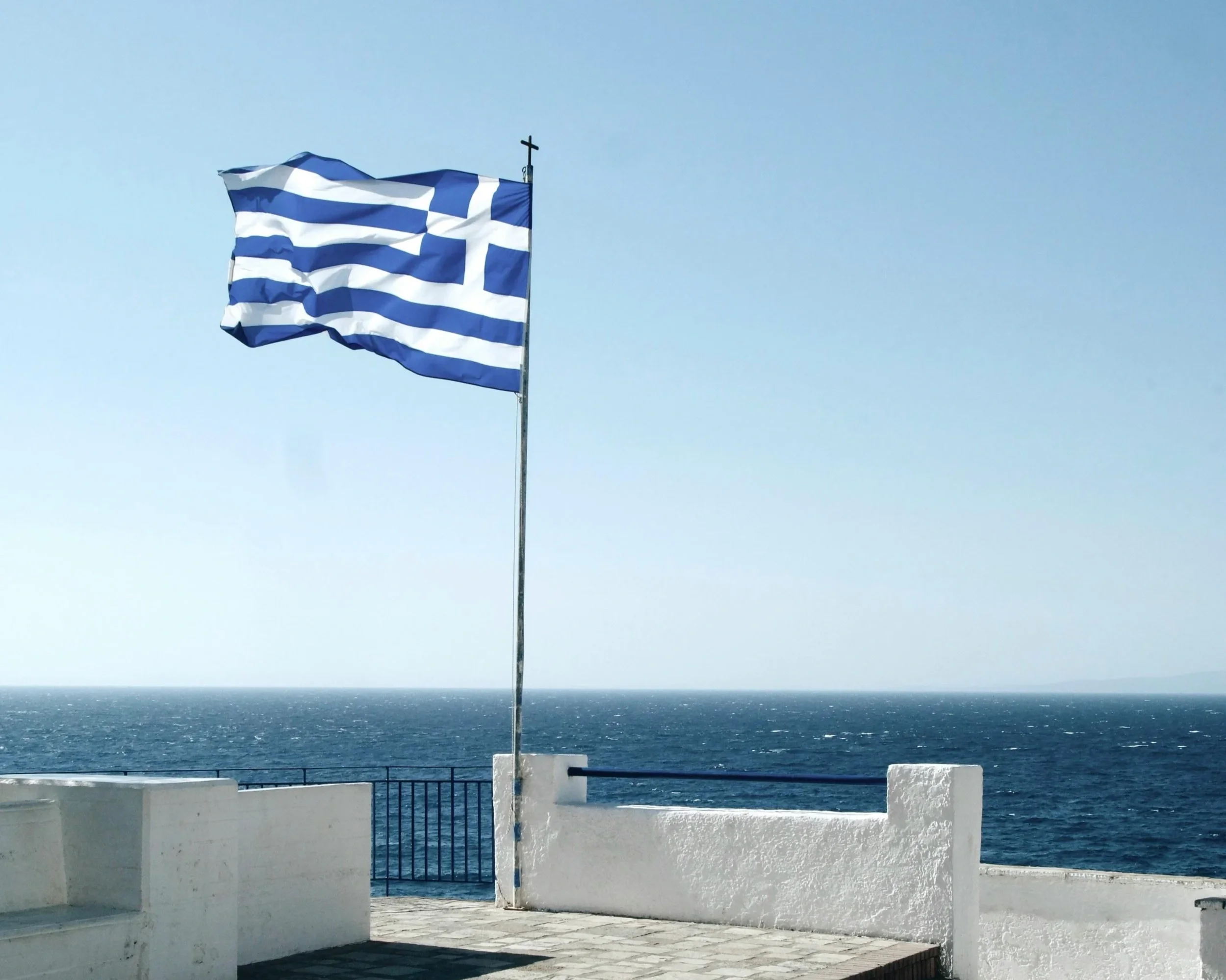 A Greek flag with blue and white stripes and a cross in the upper left corner, flying on a flagpole by the ocean under a clear blue sky, with a white wall and railings in the foreground.