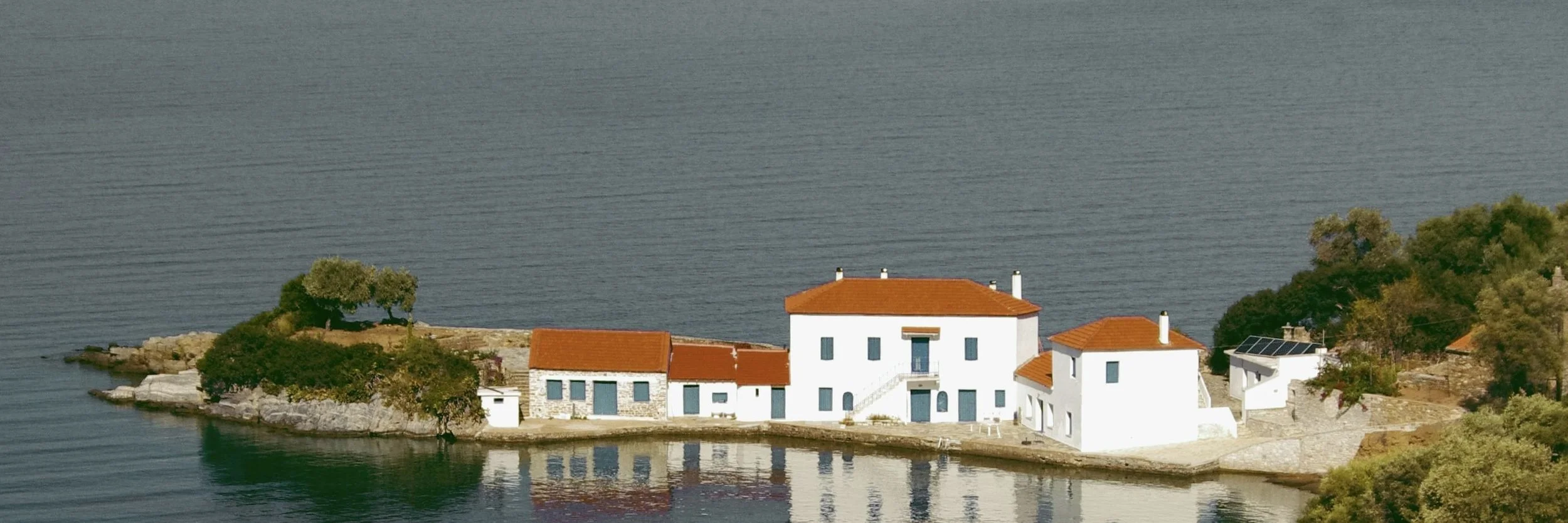 A small island with white buildings with red roofs, surrounded by water, with trees and greenery.