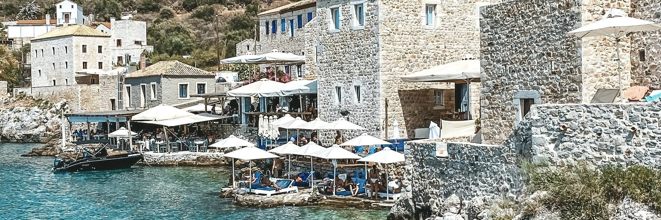A coastal scene with stone buildings, some with blue shutters, and outdoor seating areas with parasols along the water, with people lounging and enjoying the view.