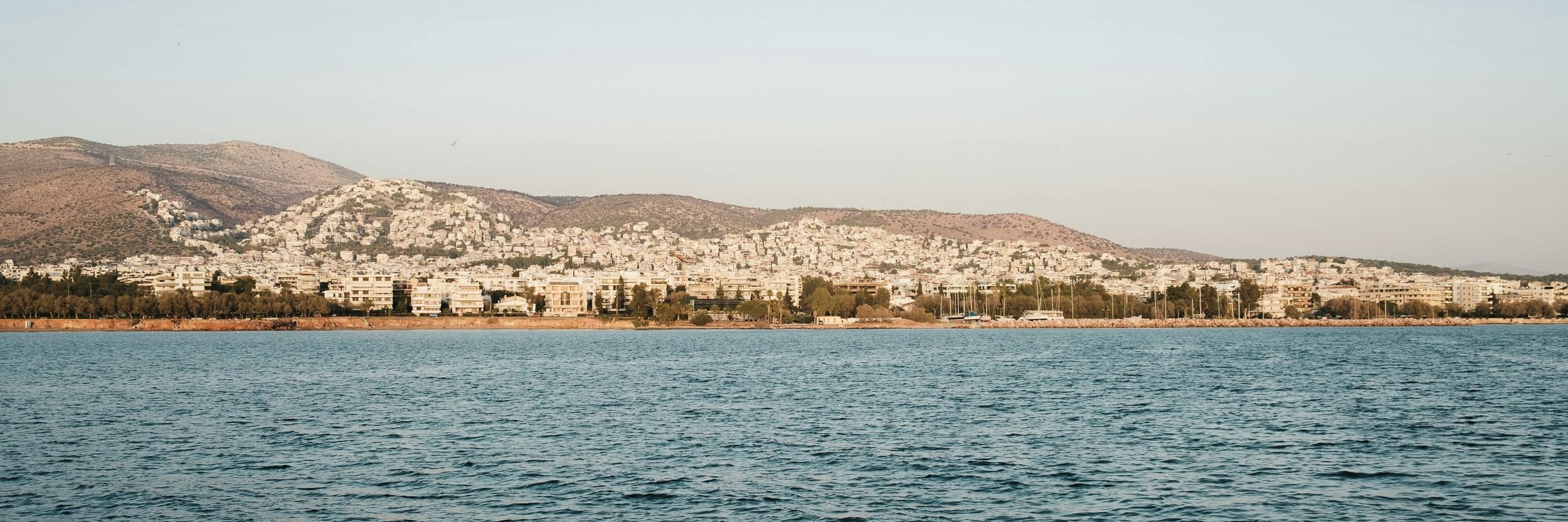 A coastal city with white buildings on a hillside, viewed across a body of water, with sailboats docked along the shore.