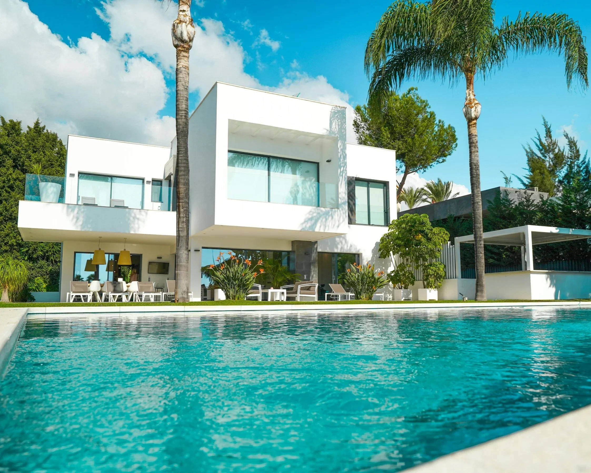 Modern white house with large glass windows and palm trees in front, swimming pool in foreground, under a blue sky with clouds, outdoor chairs and tables around the pool.