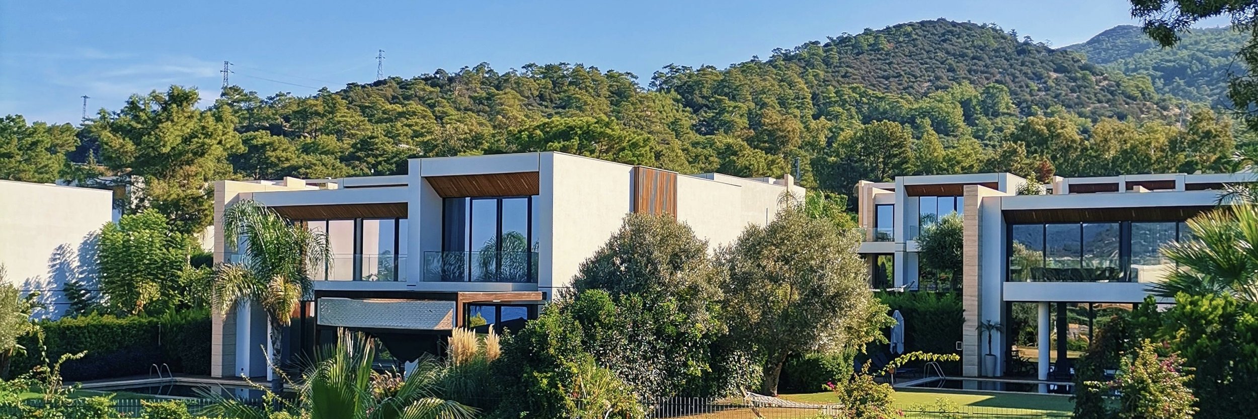 Modern white residential buildings with large glass windows and balconies, surrounded by lush greenery and trees, with a forested hillside in the background.