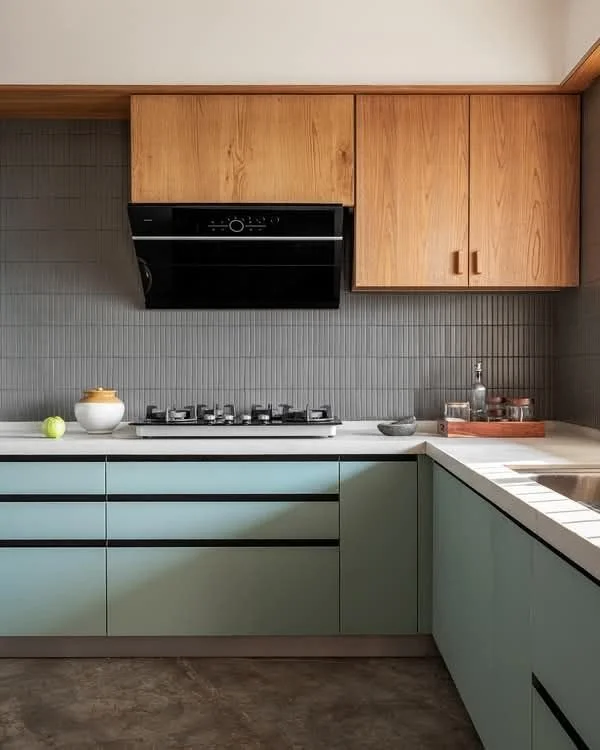 Modern kitchen with wooden upper cabinets, light green lower cabinets, gray tiled backsplash, and a white countertop. Includes a black range hood, gas stove, and small decorative items on the counter.