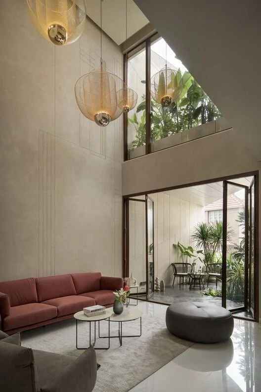 Modern living room with high ceilings, large windows, and a mix of contemporary furniture including a pink sofa, round coffee tables, and a black ottoman. There are hanging pendant lights and a small balcony or patio area with plants visible through wide glass doors.