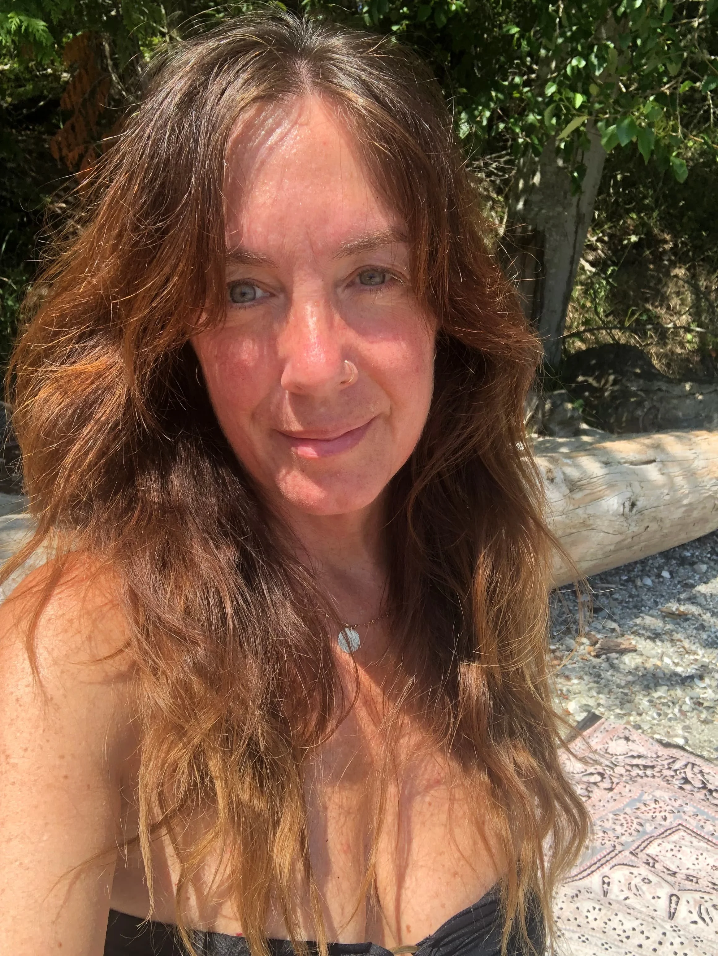 A woman with long, wavy, reddish-brown hair taking a selfie outdoors. She is illuminated by sunlight, has a light complexion, and blue eyes. She is wearing a necklace with a round pendant, a black top, and a nose ring. Behind her, there are green leafy bushes, a fallen tree trunk, and rocks on the ground.