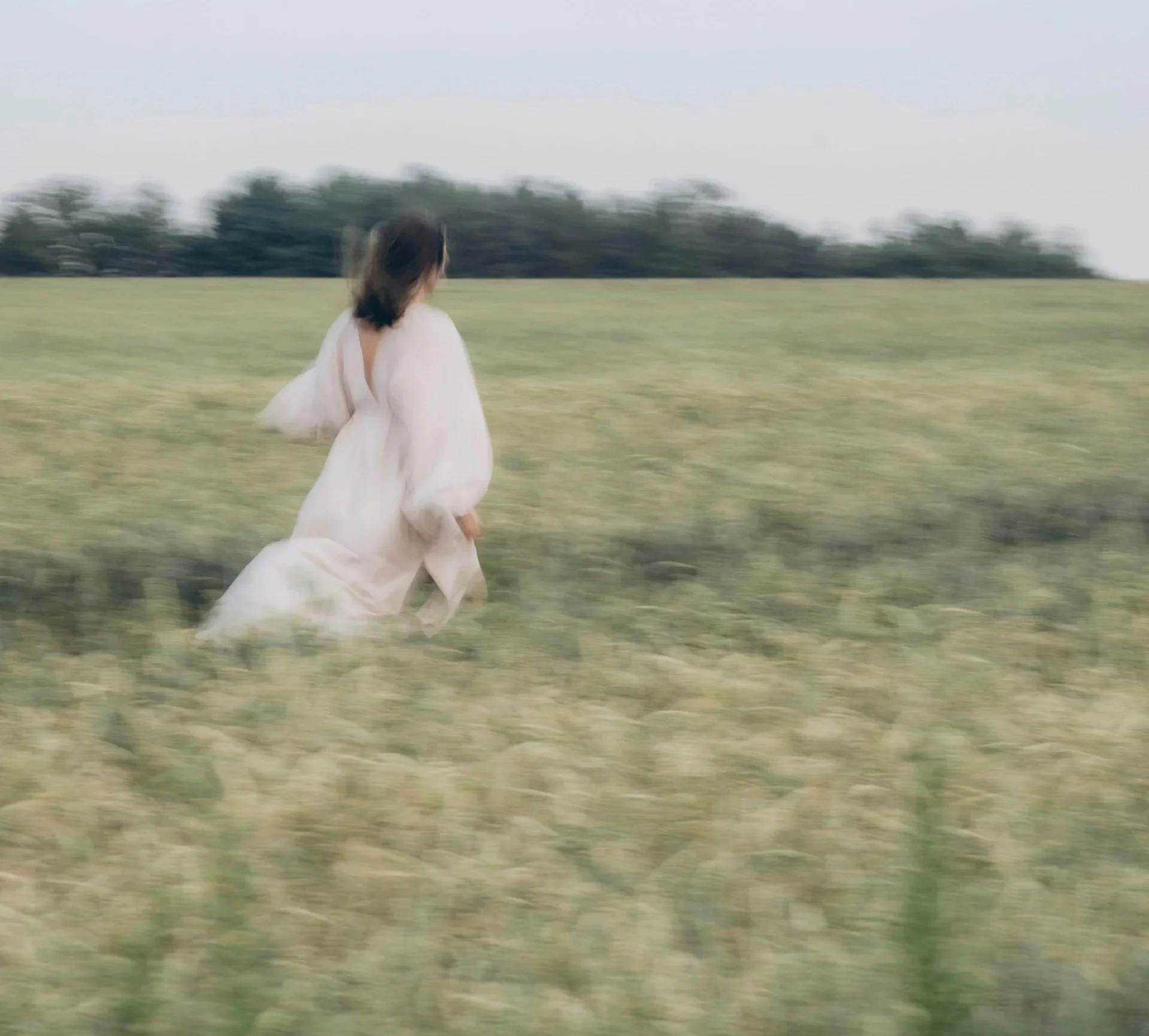 A woman in a white dress running through a green field with trees in the background.