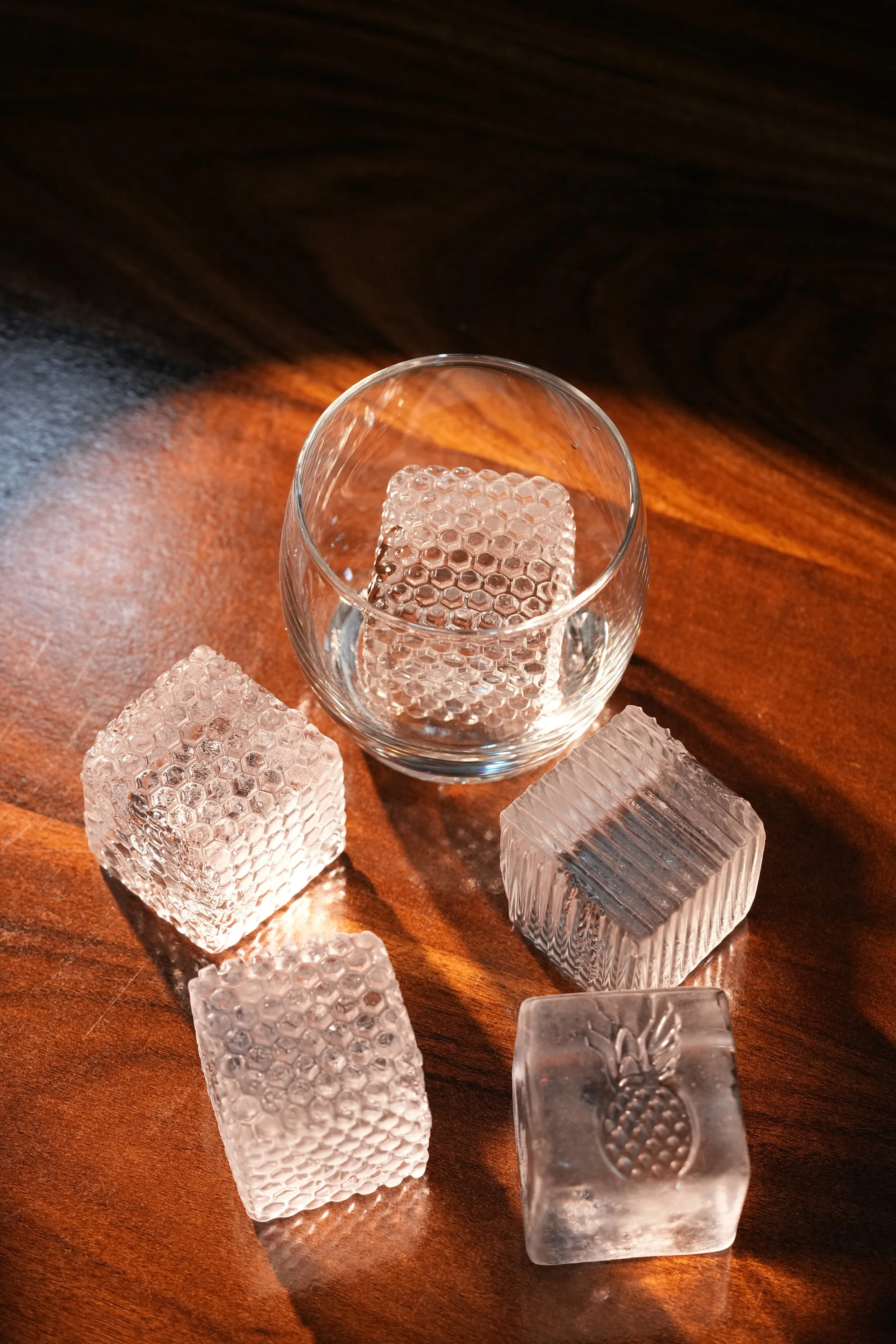A clear glass with a square ice cube inside, surrounded by five other square and rectangular ice cubes with textured surfaces, one of which has a pineapple engraving, on a wooden surface.