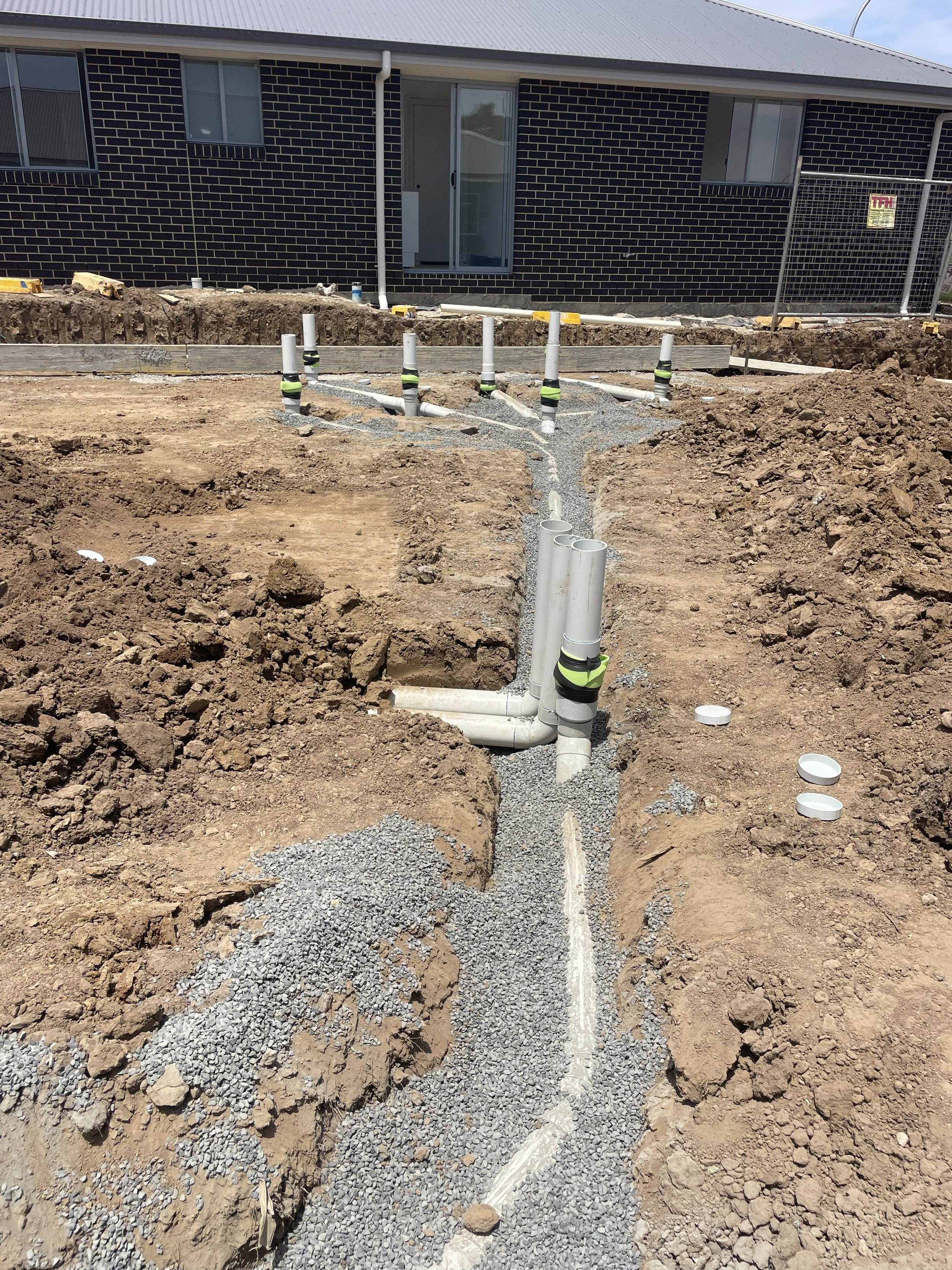 Construction site with pipes and gravel for plumbing installation in front of a residential building with black brick exterior.