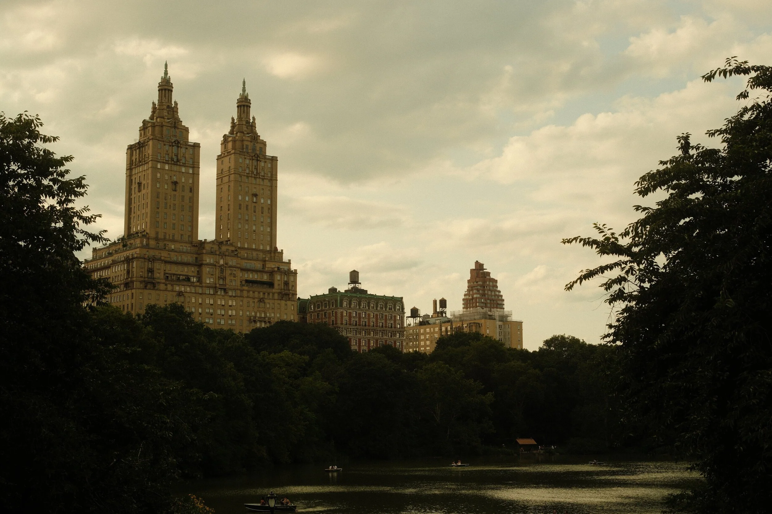 Cityscape with tall buildings seen from a park with trees and a river, boats, under a cloudy sky.
