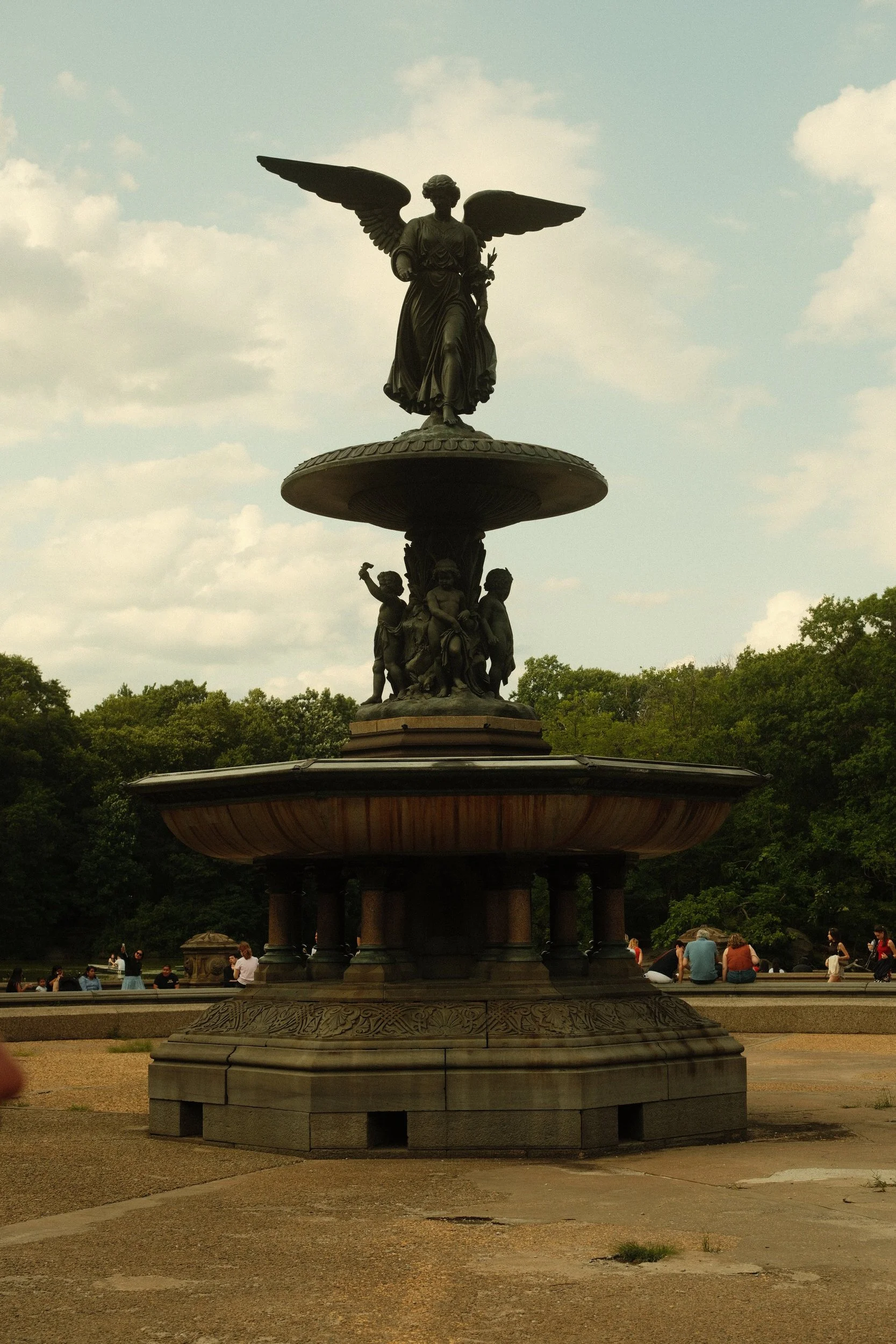 A bronze fountain featuring a statue of a winged woman holding a branch, surrounded by children, in a park with trees and visitors.