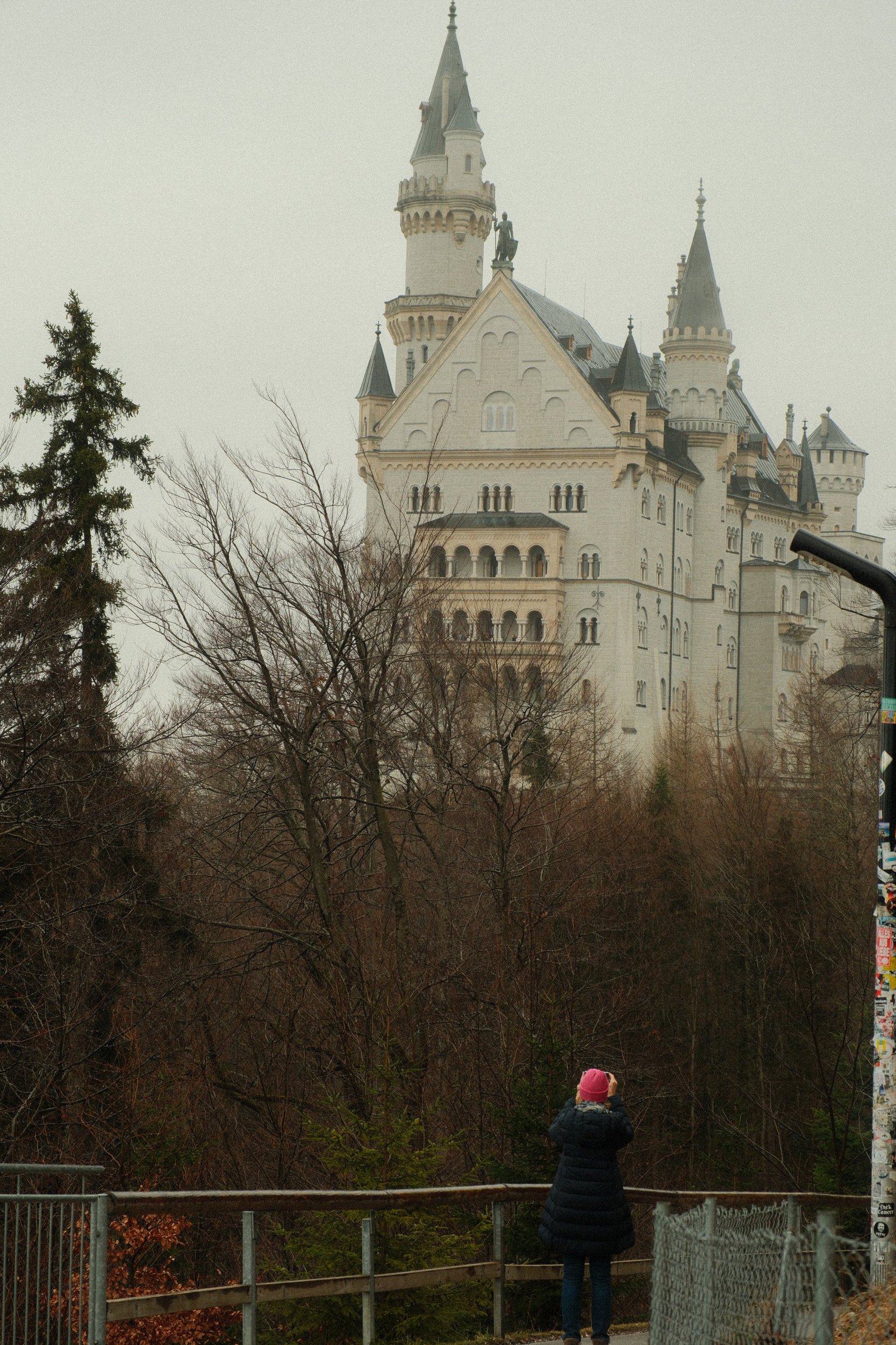 A person in a dark coat and pink hat taking a photo of a large, fairytale-style castle on a cloudy day, with leafless trees in the foreground.