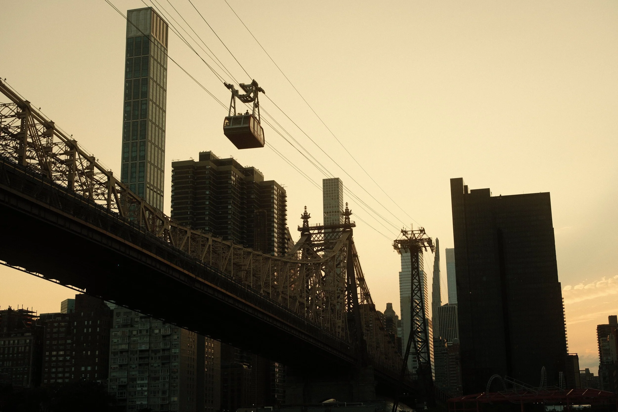View of a city skyline with tall buildings, including a tower with glass windows, and a bridge with a cable car hanging from cables above the bridge, all under a dusky sky.