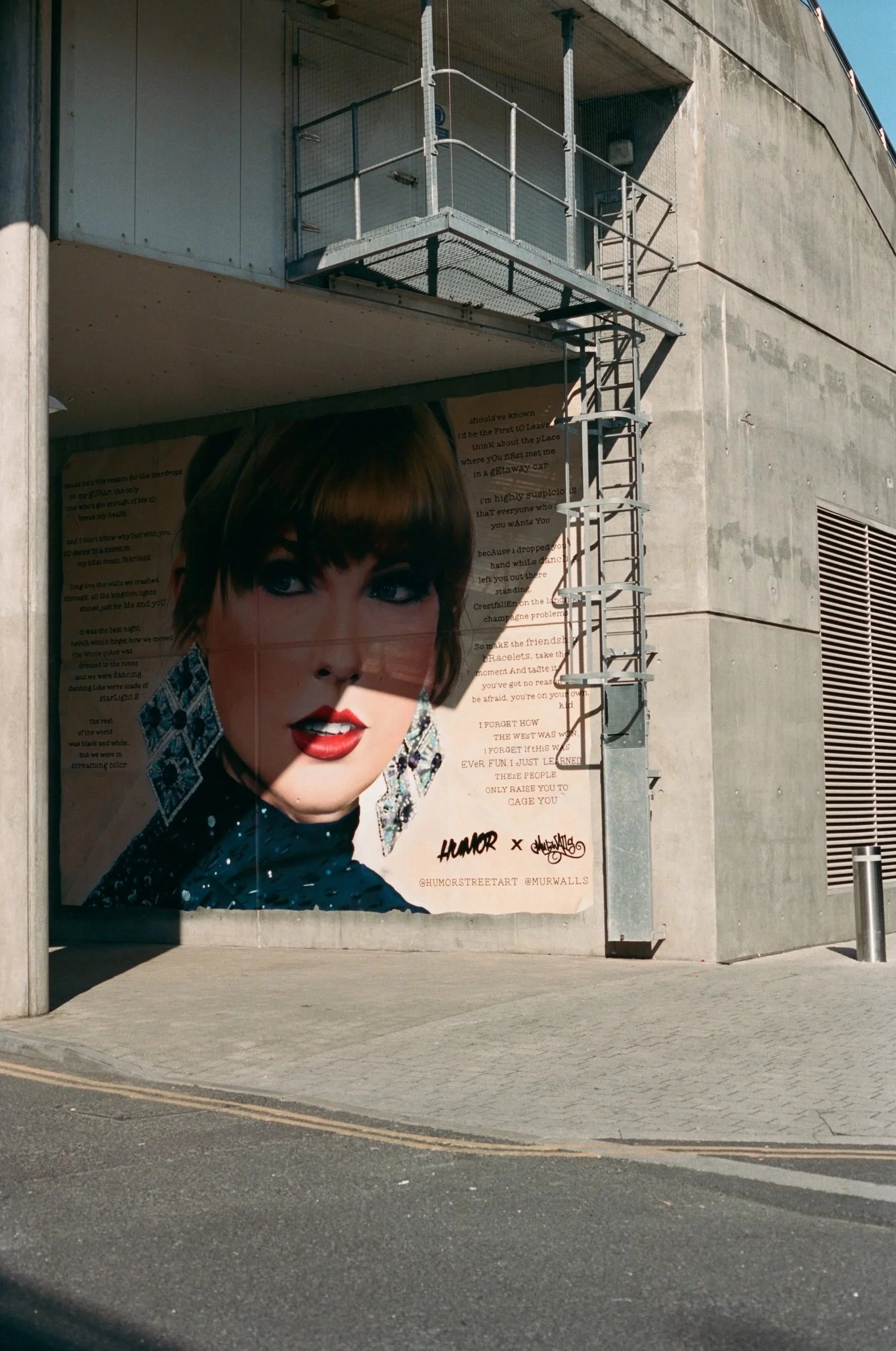 A large advertisement featuring a woman's face with makeup and earrings on a concrete wall, with text and signatures at the bottom, and a fire escape staircase attached to the building.