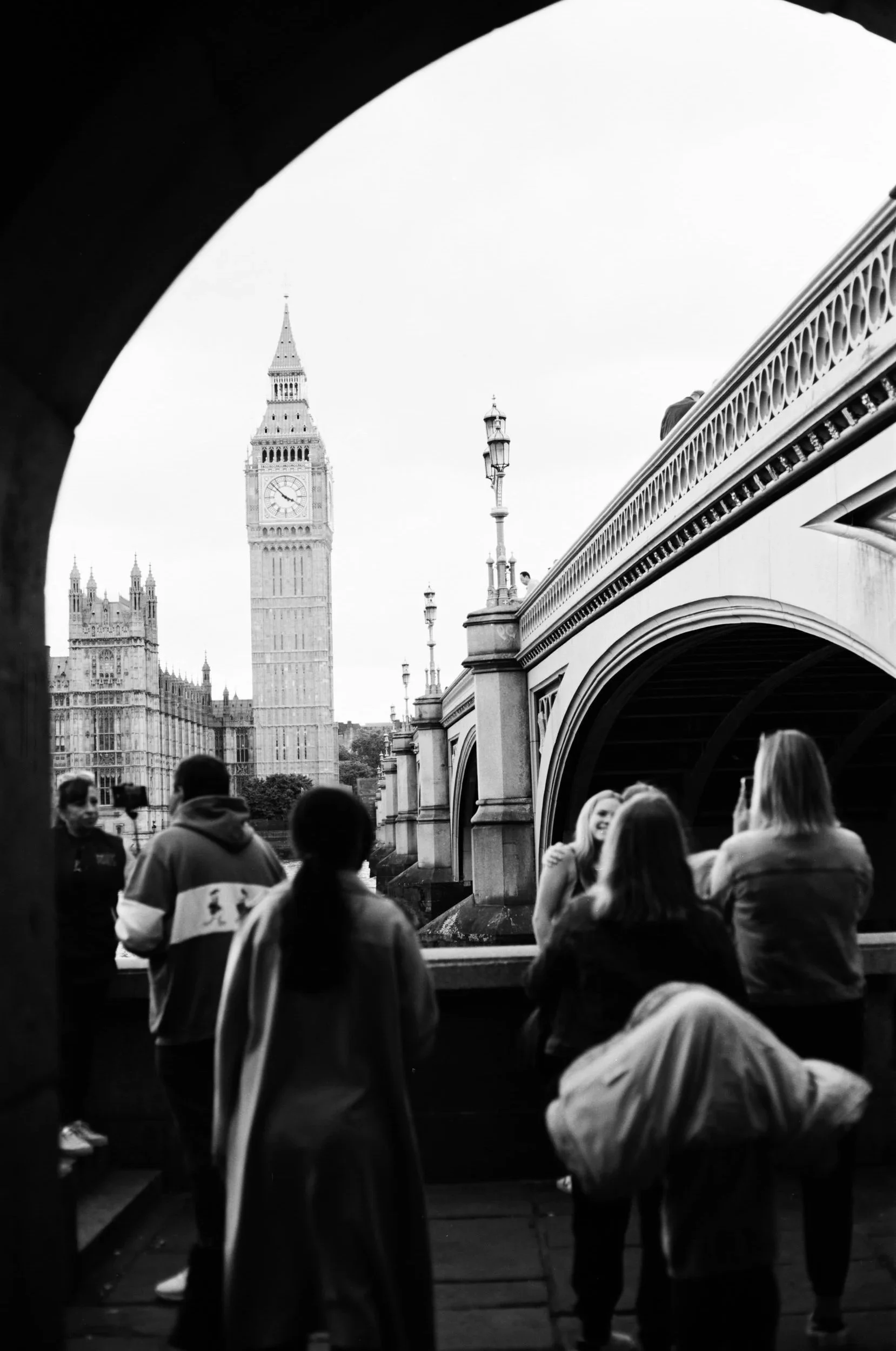 Black and white photo of Big Ben and the Houses of Parliament in London, viewed through an archway, with several people standing on a bridge in the foreground.