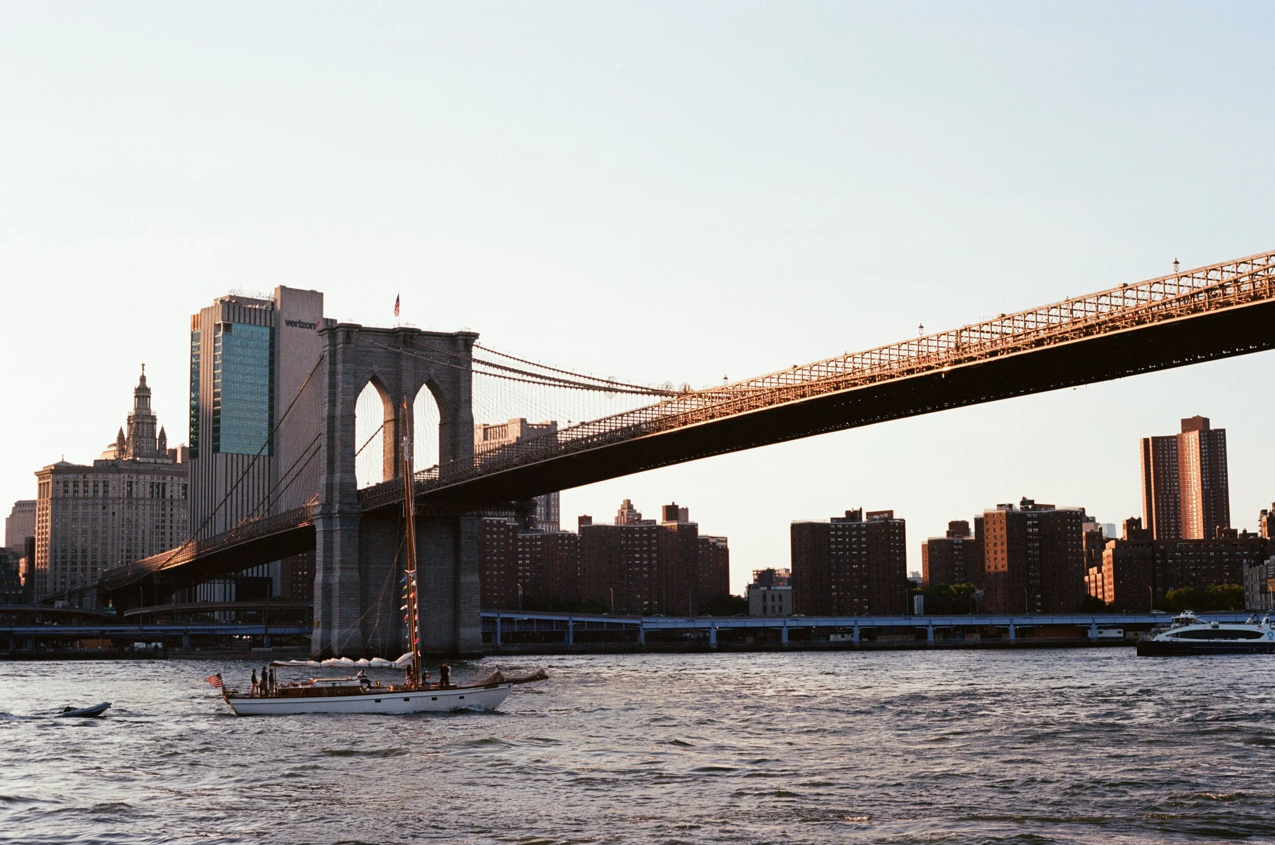 View of the Brooklyn Bridge in New York City with skyscrapers in the background and a sailboat on the water in the foreground during sunset.