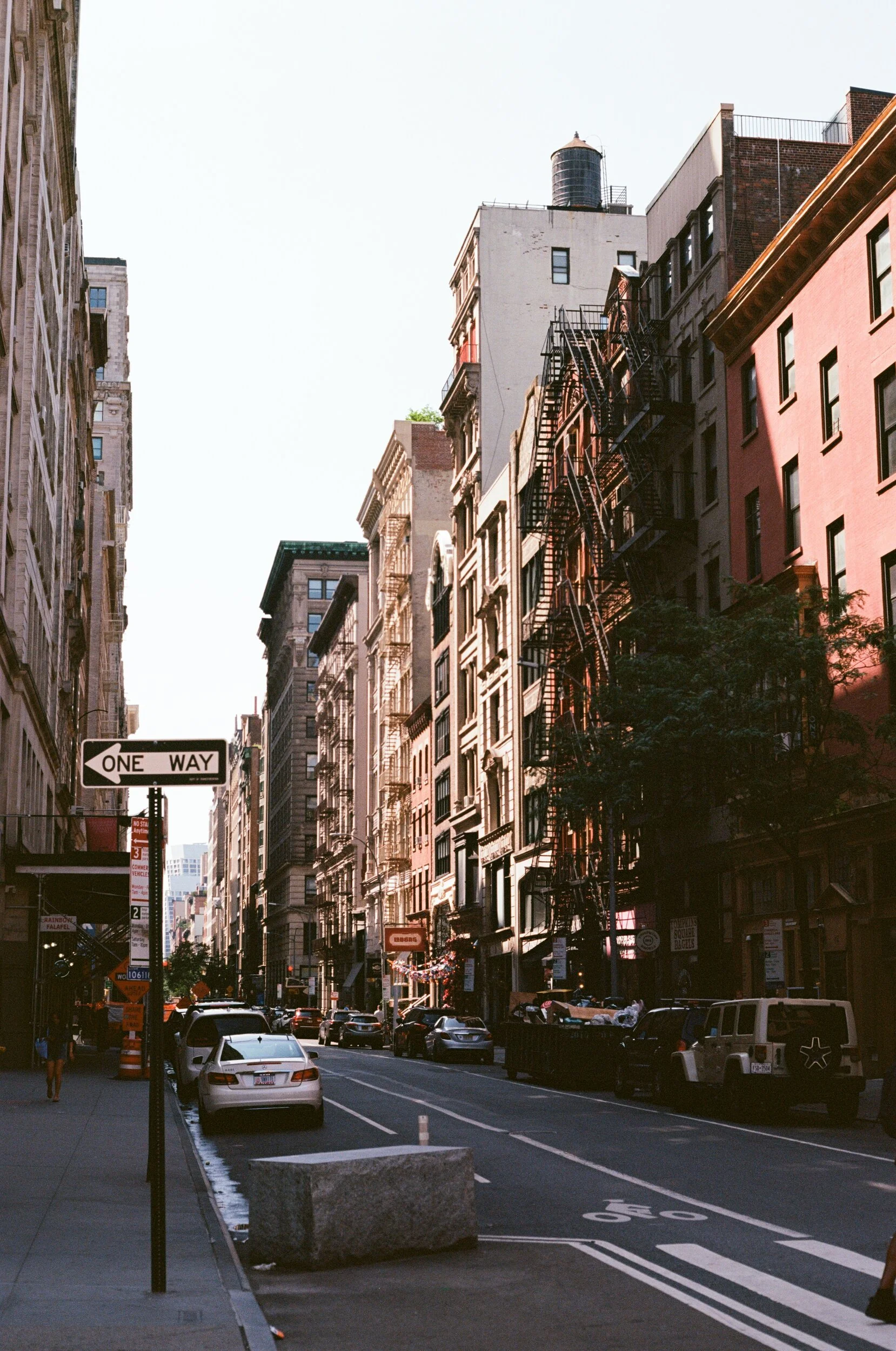 City street scene with tall buildings, parked cars, and a one-way traffic sign, during the daytime.