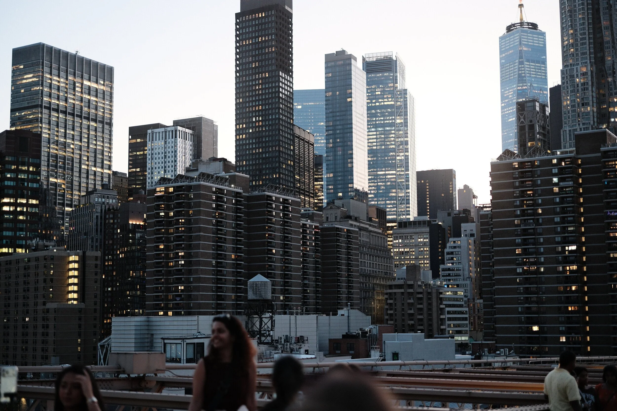 City skyline with tall buildings and skyscrapers at dusk, with people gathered on a rooftop or balcony in the foreground.