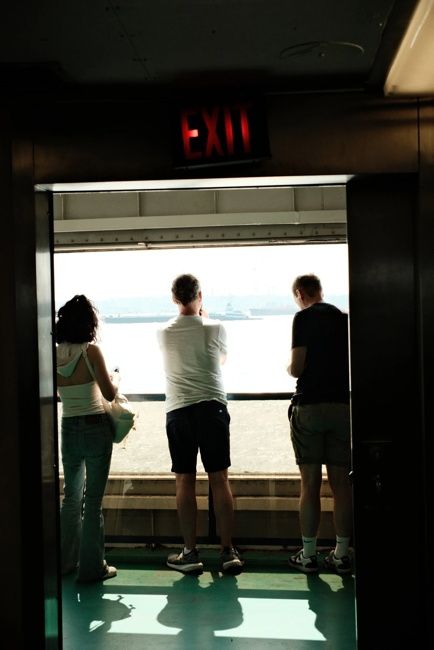 Four people looking out from a ferry window at the water and city skyline, with an illuminated exit sign above.