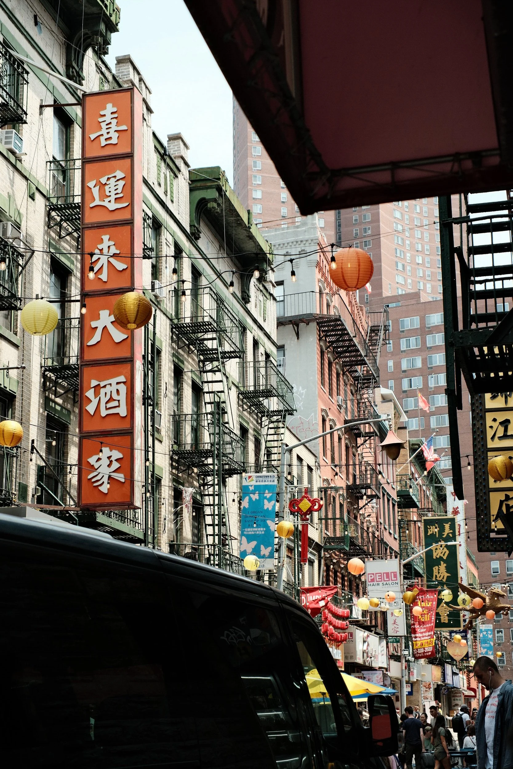 Street scene in Chinatown, with colorful signs in Chinese characters, hanging lanterns, and many pedestrians walking on the street. Tall buildings and a mix of shops and restaurants are visible.
