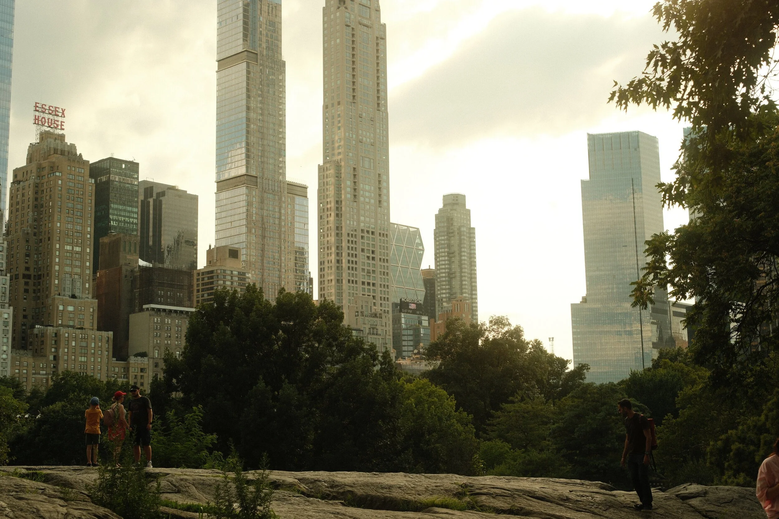 People walking in a park with a city skyline of tall buildings in the background, including skyscrapers and the Essex House sign, during daylight.