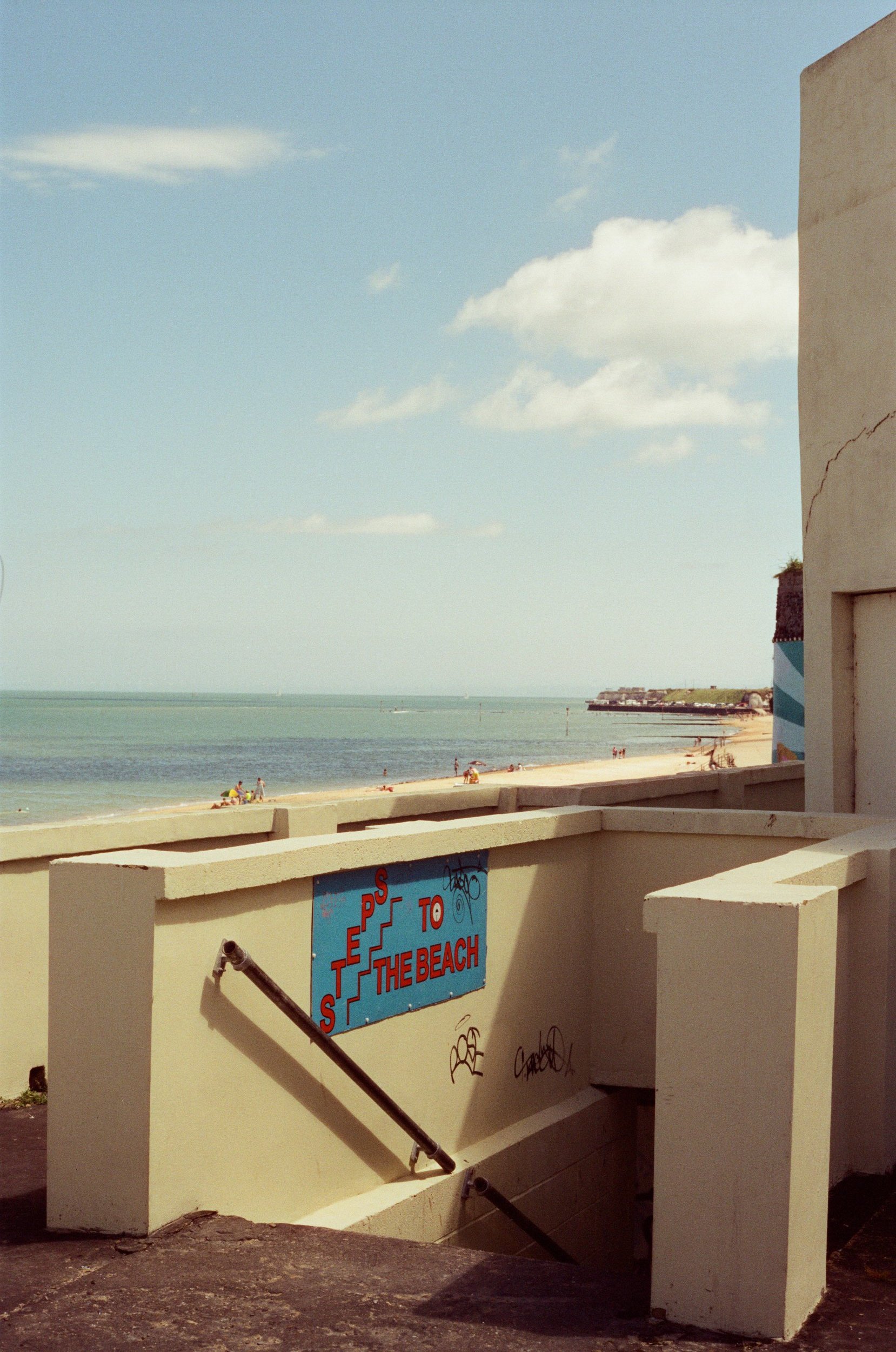 View of a beach with sand, water, and a clear sky, seen from the stairs of a building. There are people relaxing on the beach, and a sign on the wall indicates 'Steps to the Beach'.