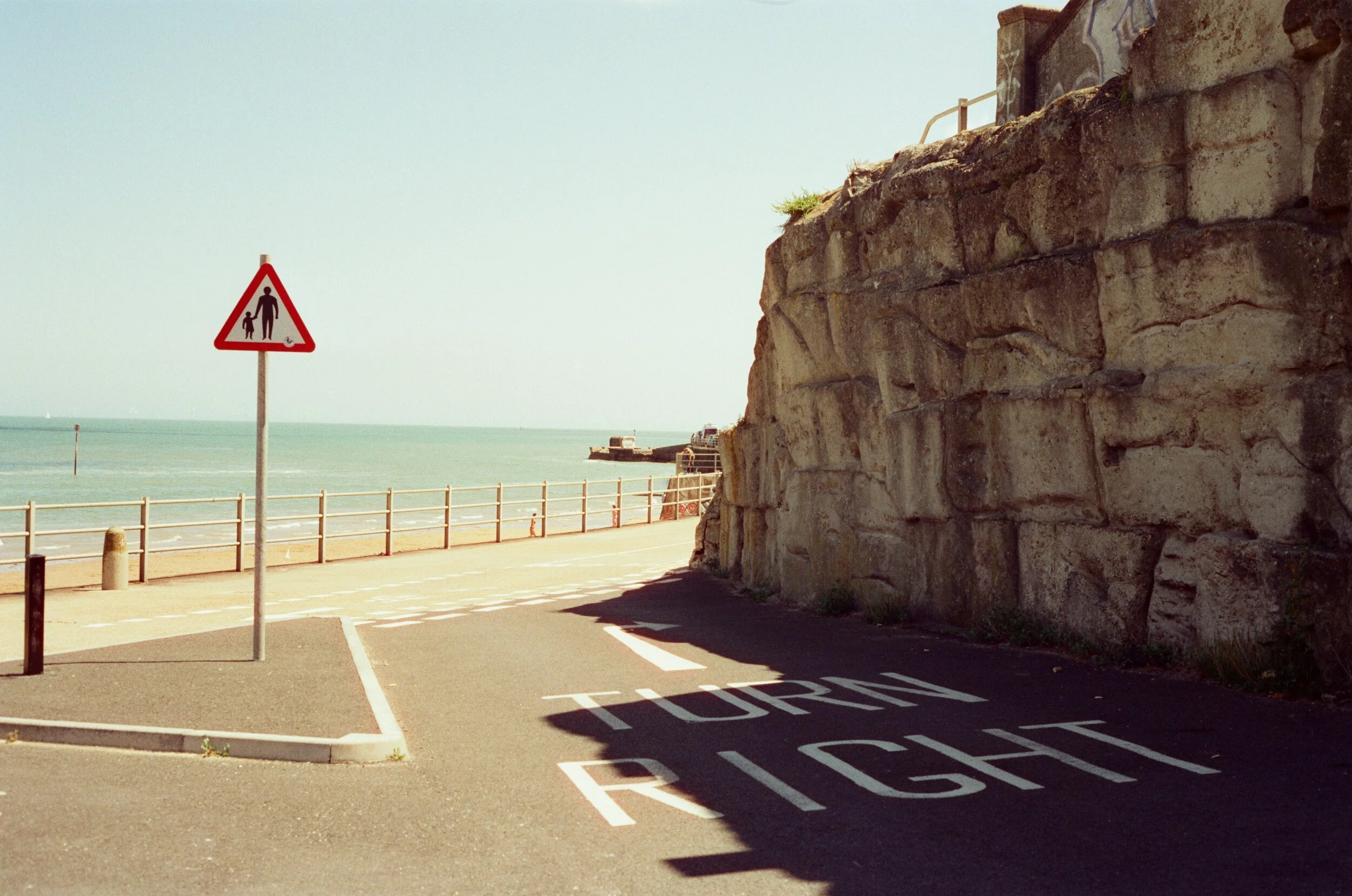 A beachside parking lot with a sign indicating pedestrian crossing and the words 'TOURISM RIGHT' painted on the ground. There is a stone wall on the right and a railing separating the parking area from the beach. The ocean is visible in the backgroun