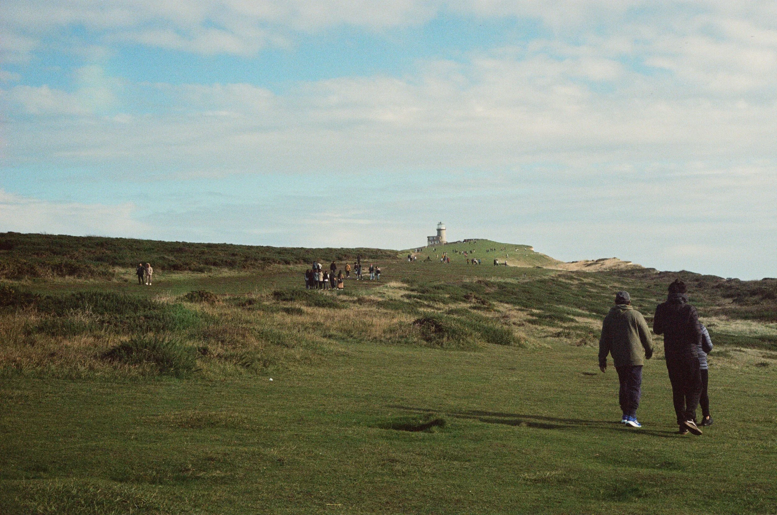 People walking on a grassy hill near a lighthouse in the distance with a partly cloudy sky.