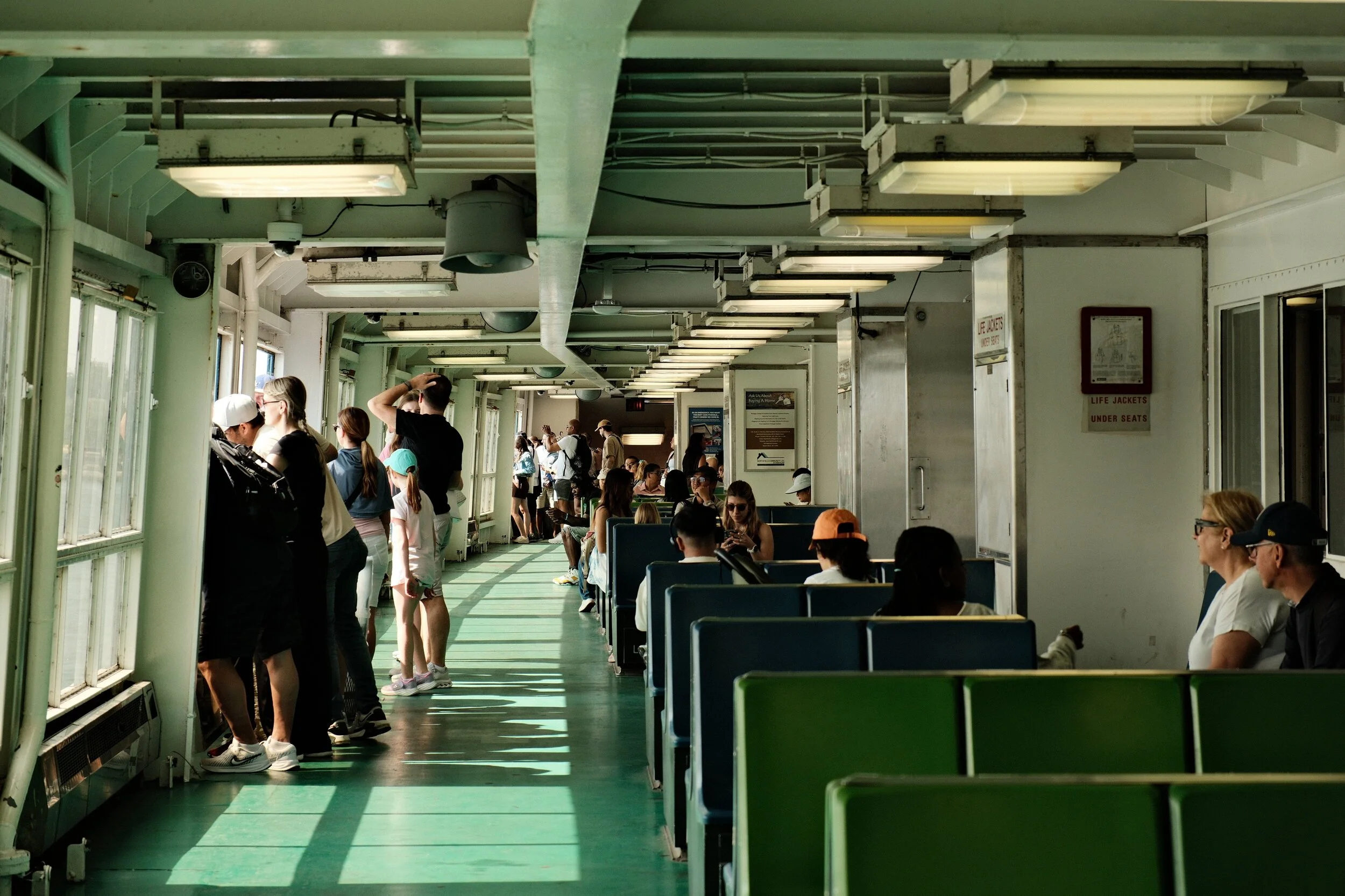 Passengers seated and standing inside a ferry or boat with large windows and green flooring, under overhead lighting.