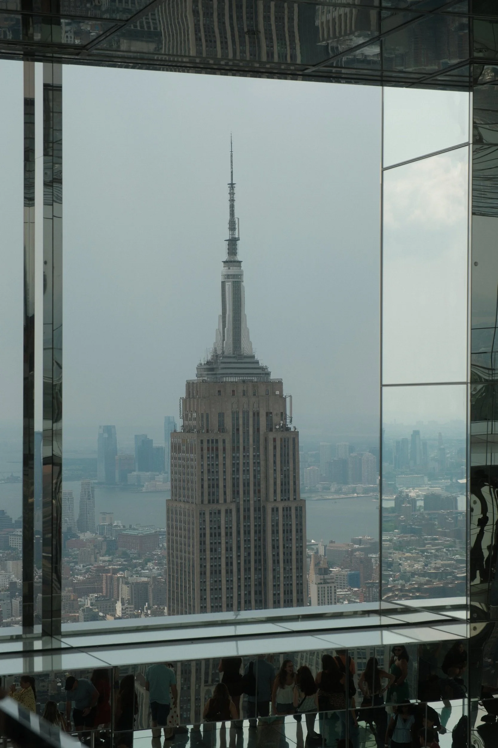 View of Empire State Building through floor-to-ceiling glass windows in a tall building, with a group of people visible inside.