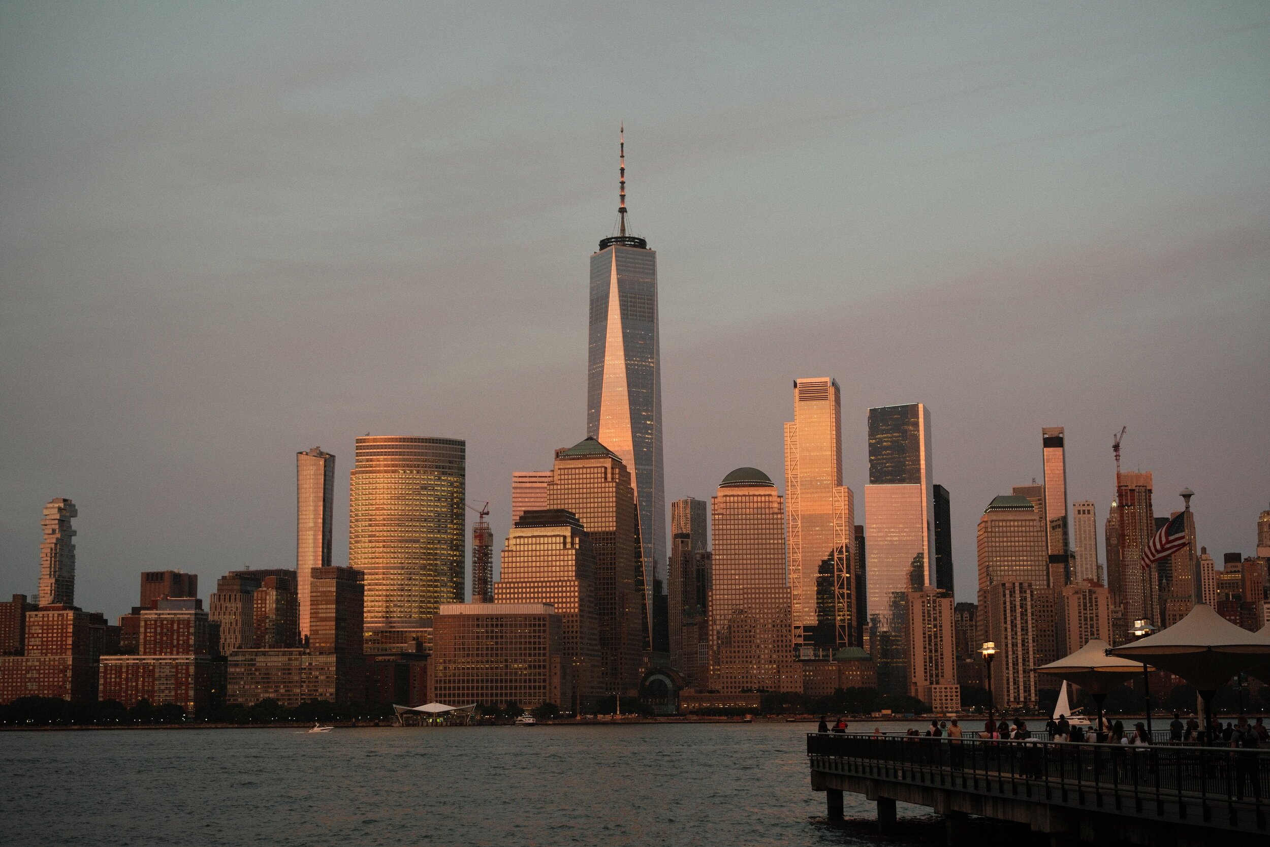 Sunset view of Manhattan skyline, featuring One World Trade Center, from waterfront with people near the railing and American flags.