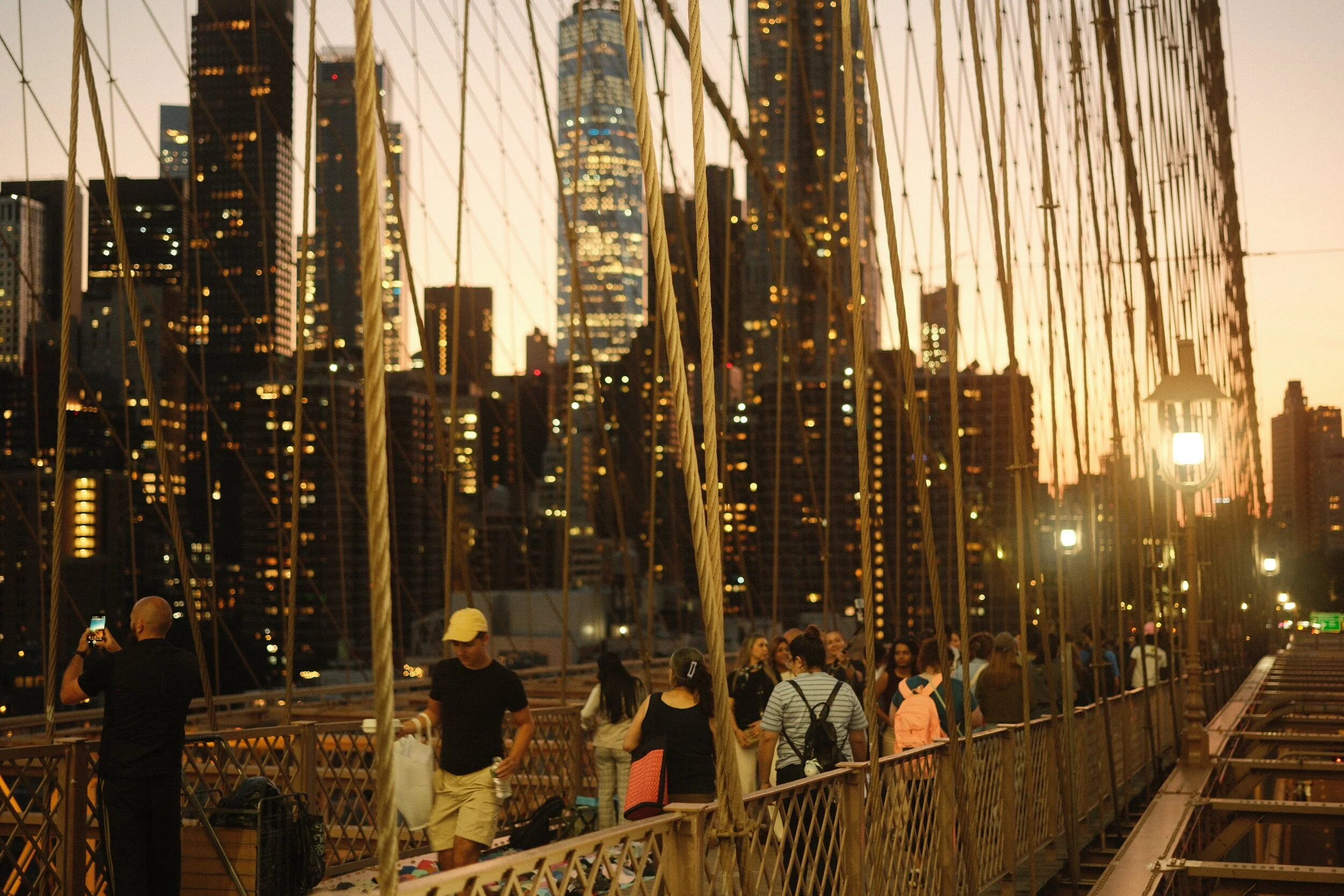 People walking on a suspension bridge at sunset with the city skyline in the background.