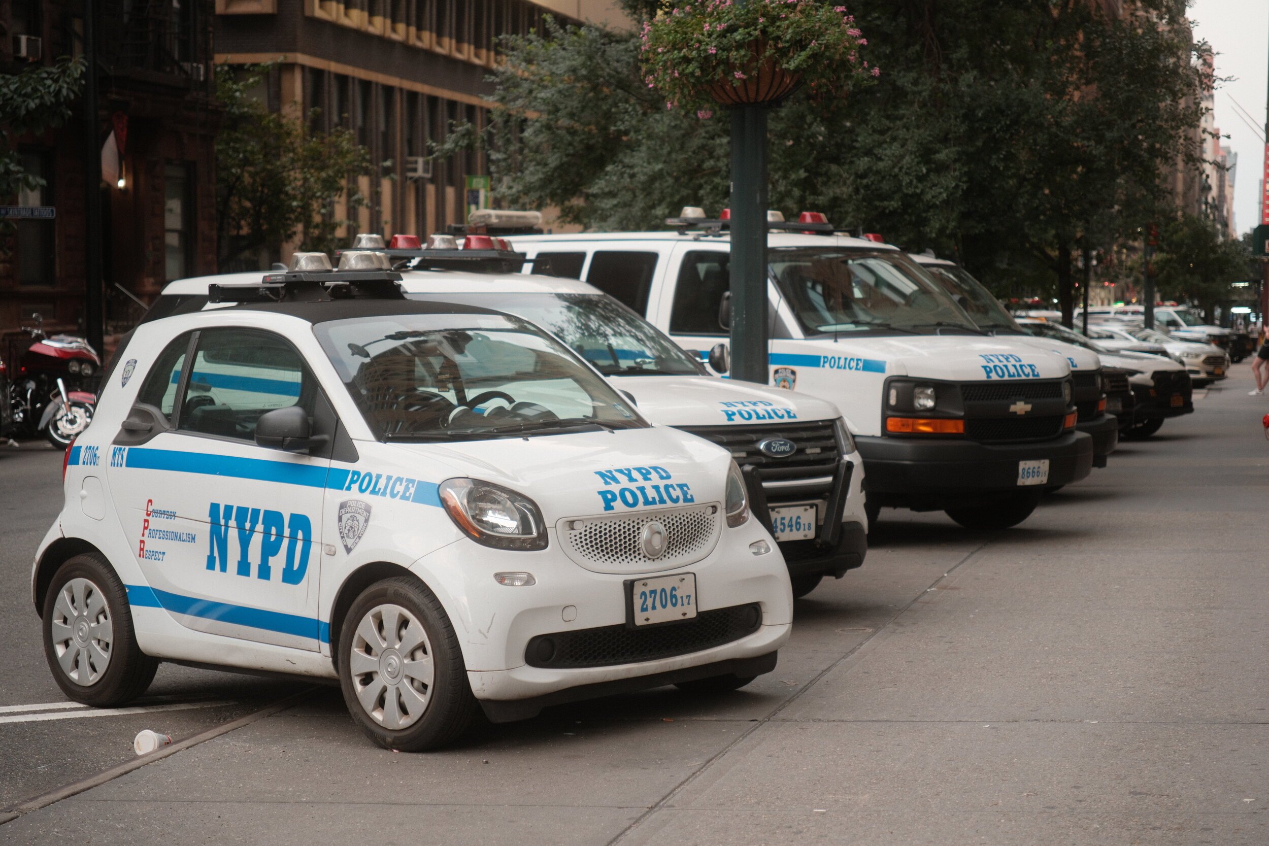 New York Police Department patrol cars parked along the street with trees and buildings in the background.