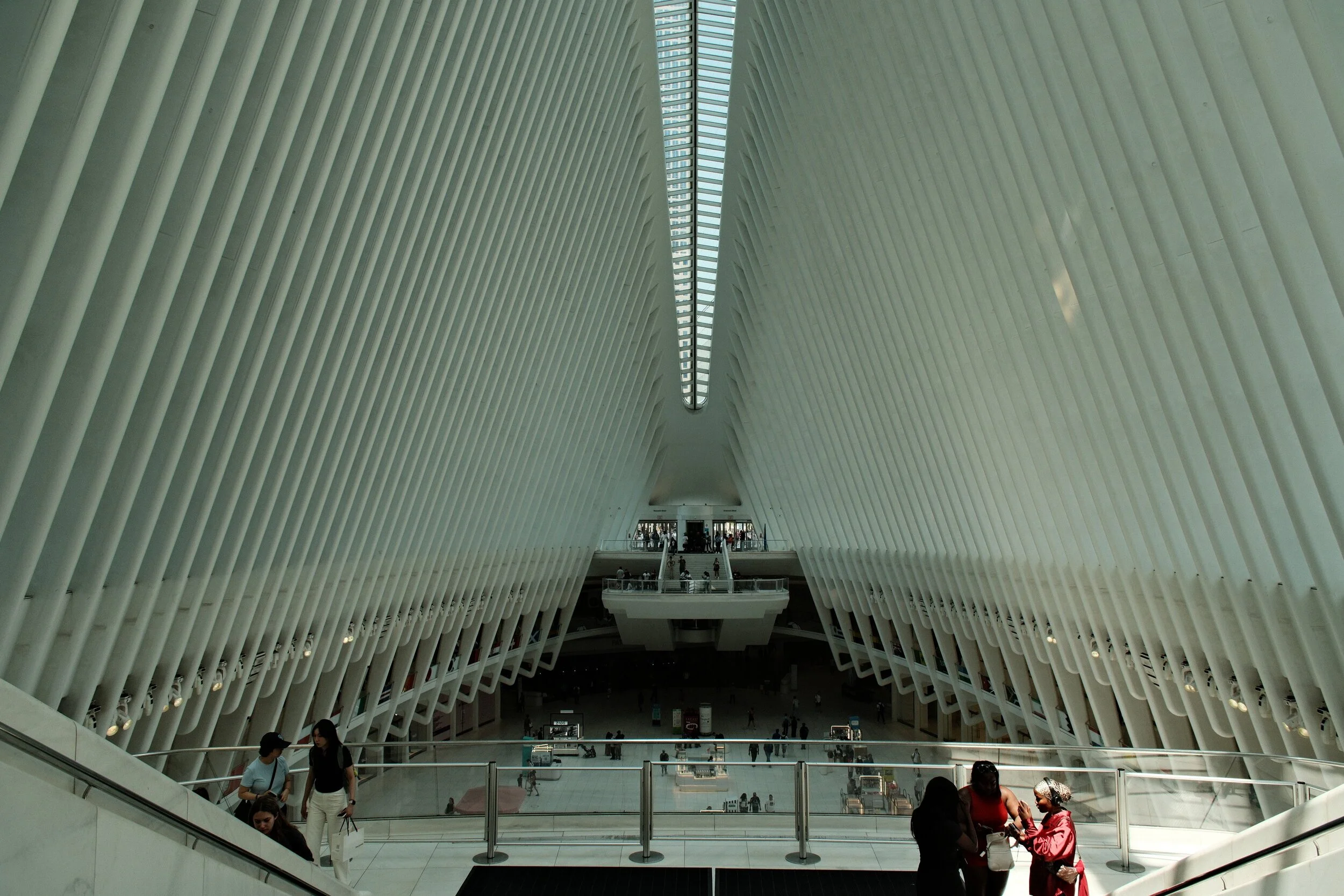 Interior of a modern airport terminal with tall white ribbed walls and a glass ceiling, showing people walking and sitting.