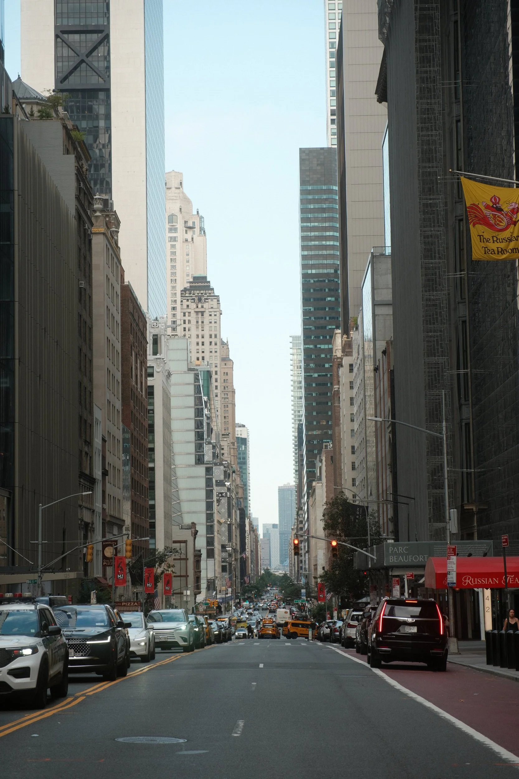 A busy city street with multiple cars and taxis, tall skyscrapers on both sides, and traffic lights visible in the foreground. Pedestrians are walking along the sidewalks, and there are signs and banners hanging from buildings.