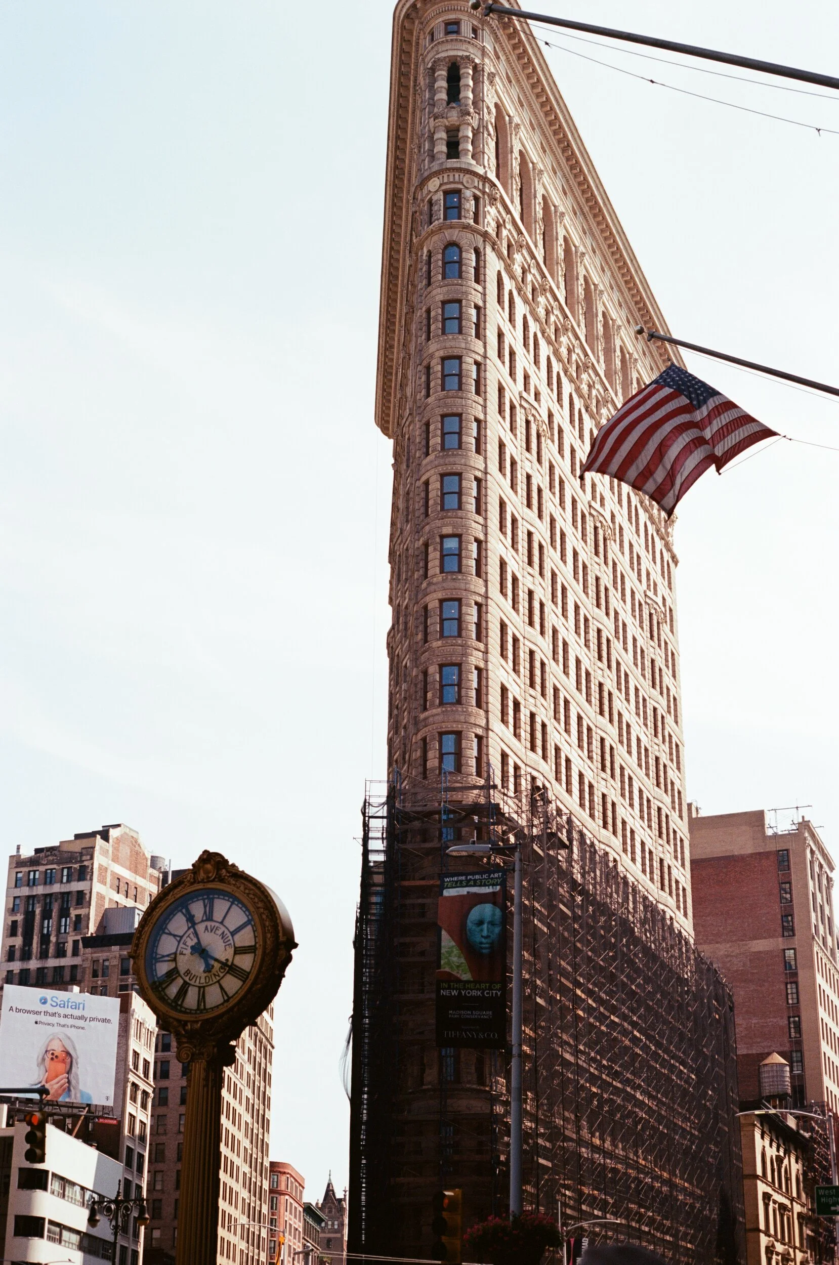 The image shows the Flatiron Building in New York City with a US flag hanging from a pole. There is a large clock on a pole with a sign indicating it is the 50th Avenue Building. The photo captures surrounding buildings and a bright sky.