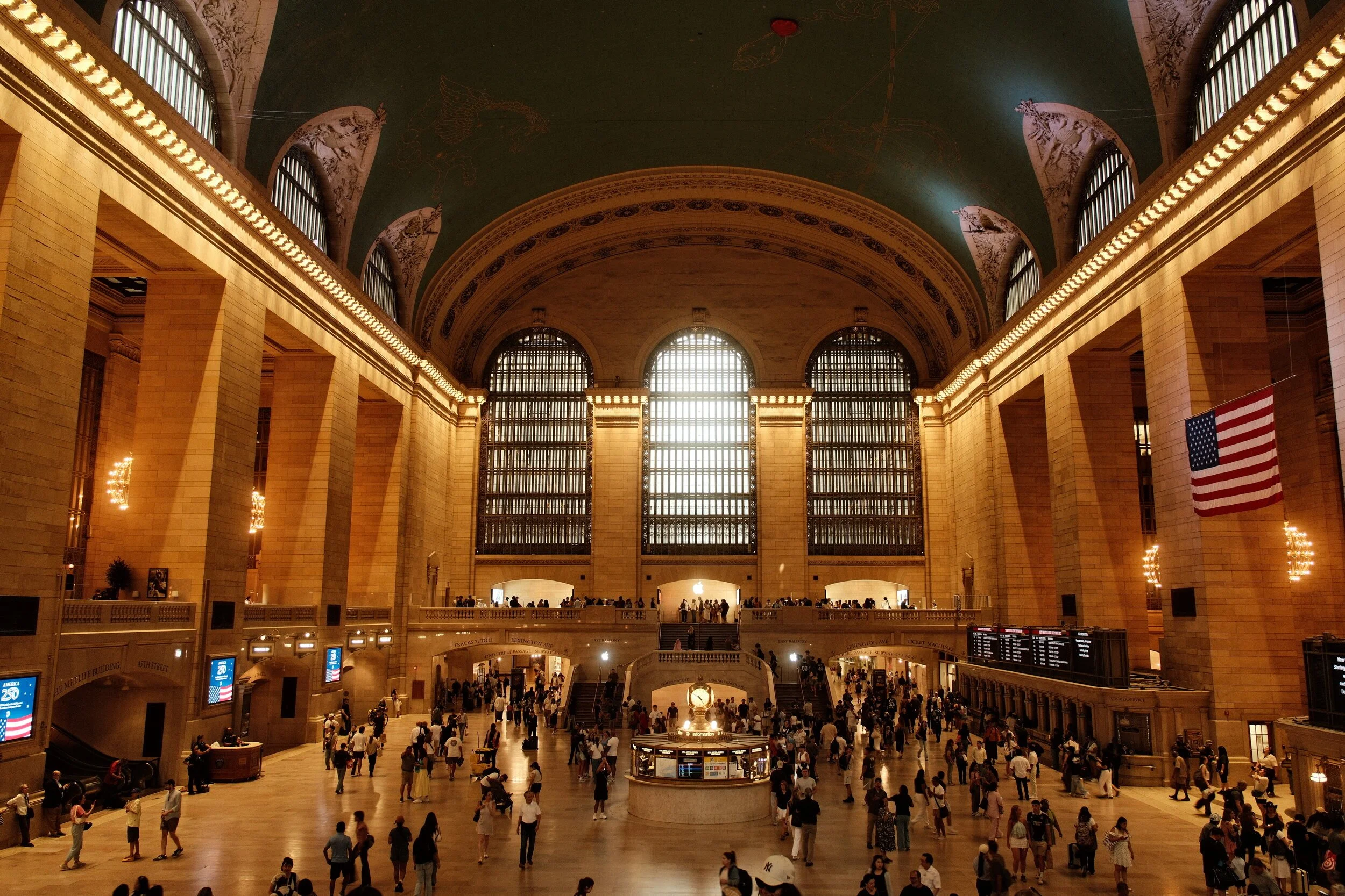 Grand Central Terminal busy interior with large arched windows, high vaulted ceiling with celestial murals, and crowds of people walking and standing.