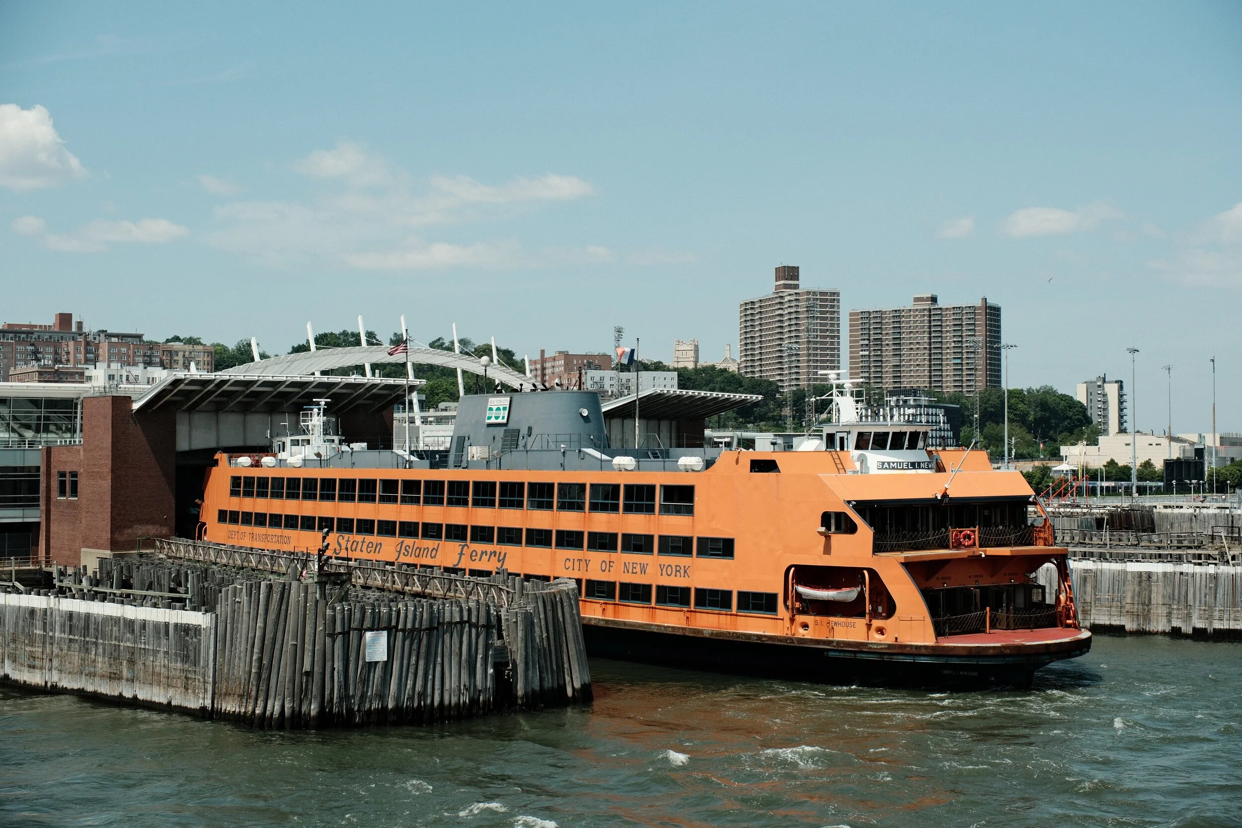 Orange Staten Island Ferry boat docked at a pier with buildings and a blue sky in the background.