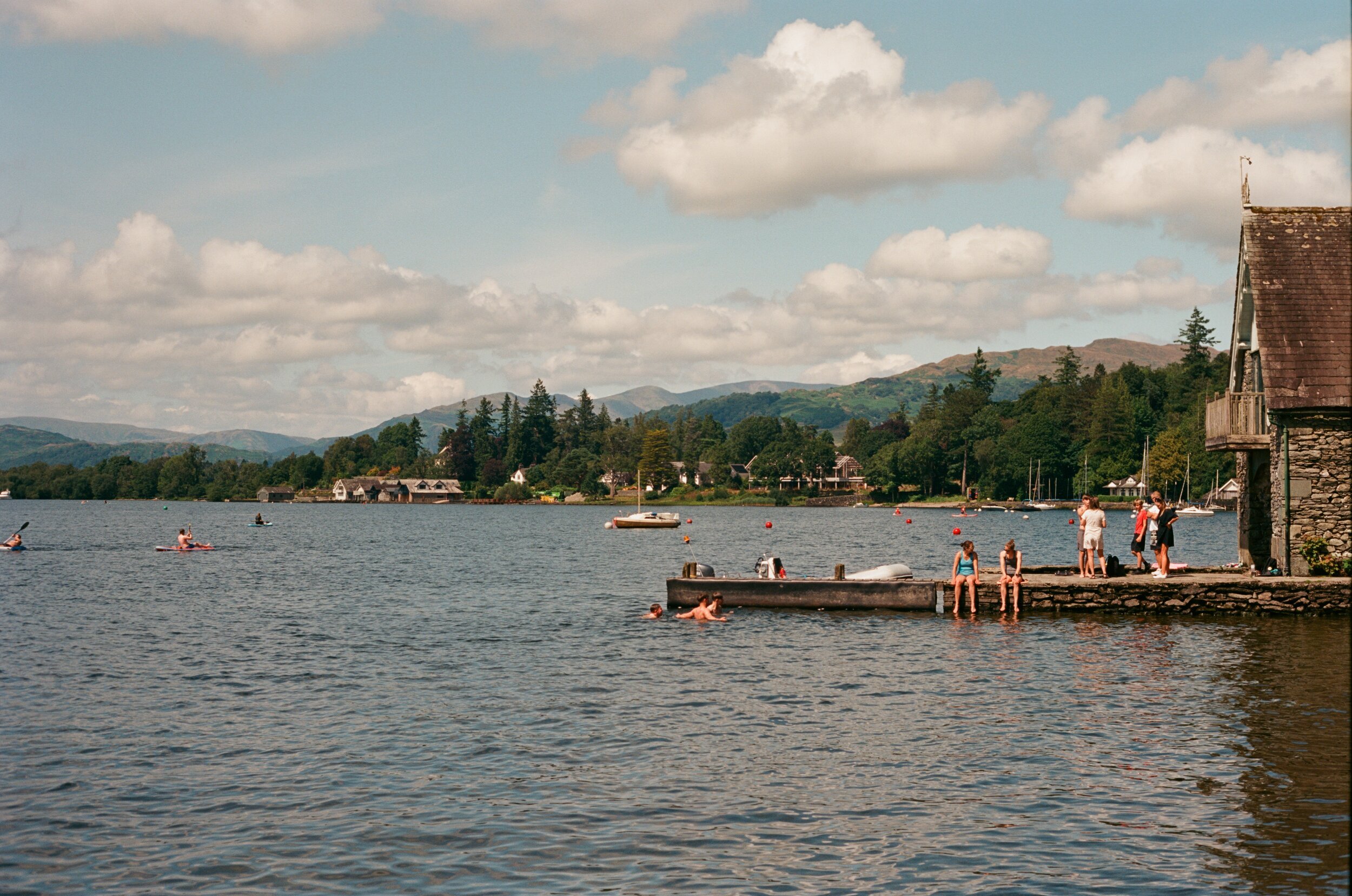 People swimming and sitting at a lakeside dock on a sunny day with a house and mountains in the background.