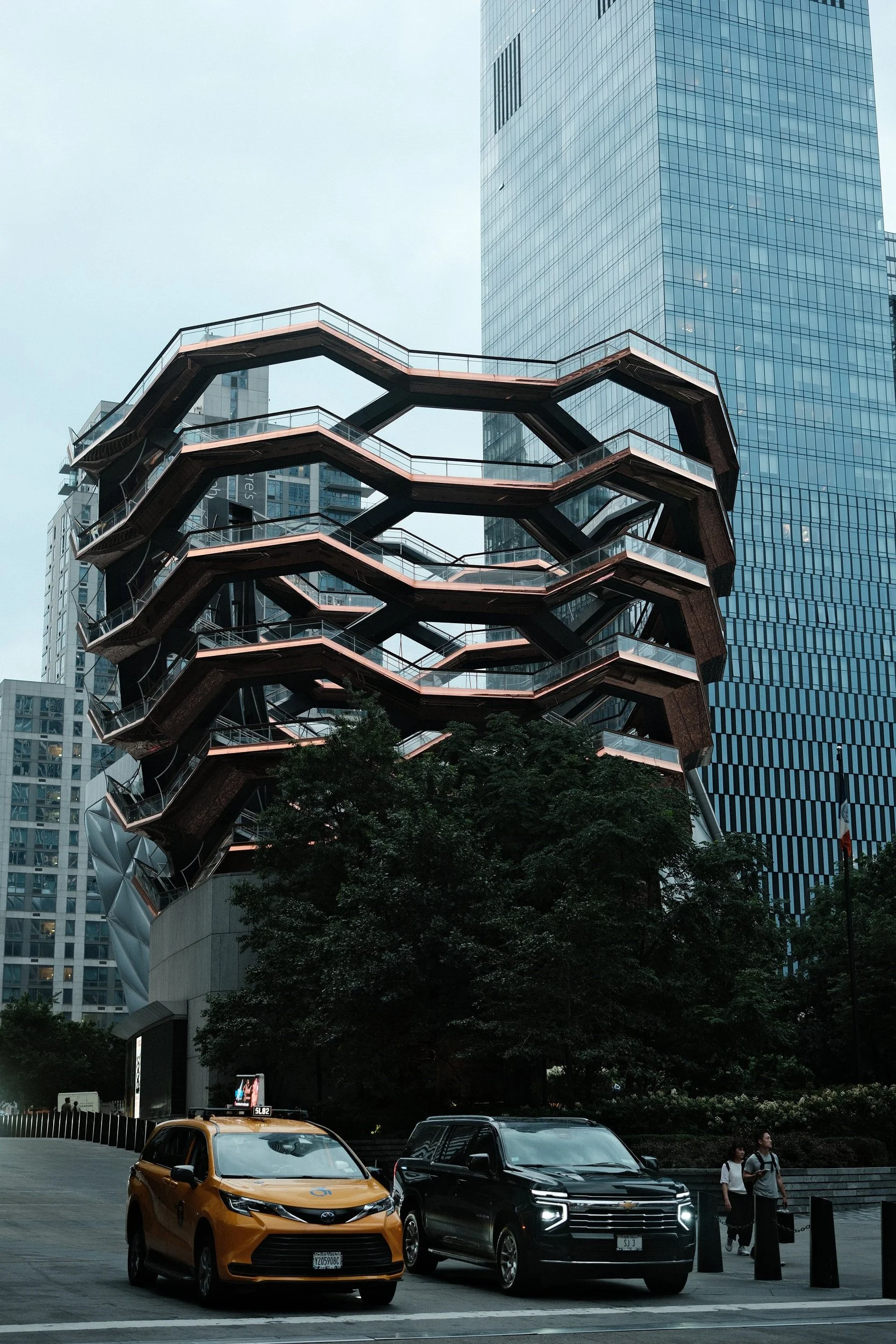 City street scene with parked cars, pedestrians, and modern high-rise buildings, including the Vessel sculpture in the background.