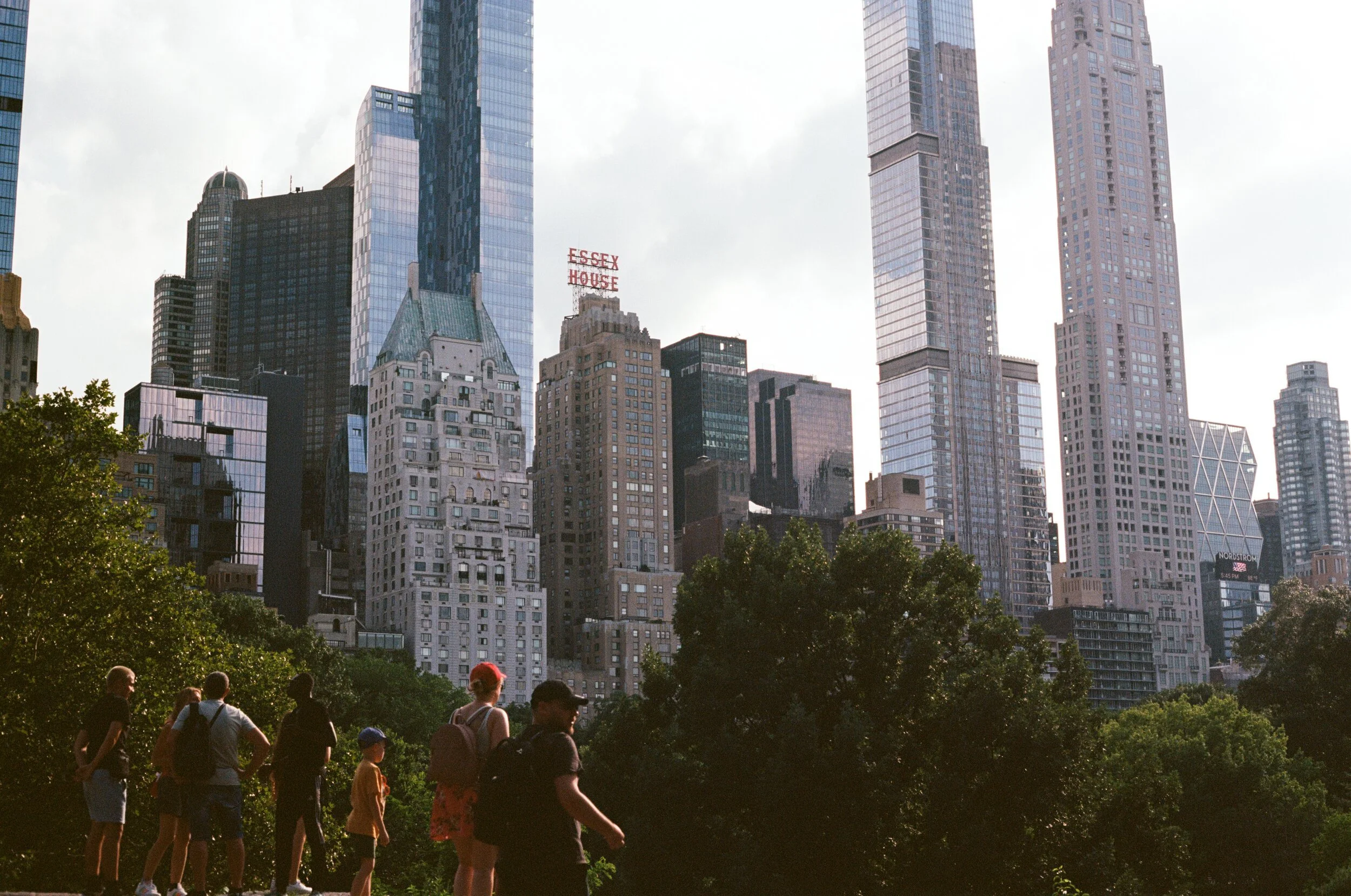 People walking in a park with tall skyscrapers in the background, including buildings with reflective glass facades and a sign that says 'Essex House' in red letters.