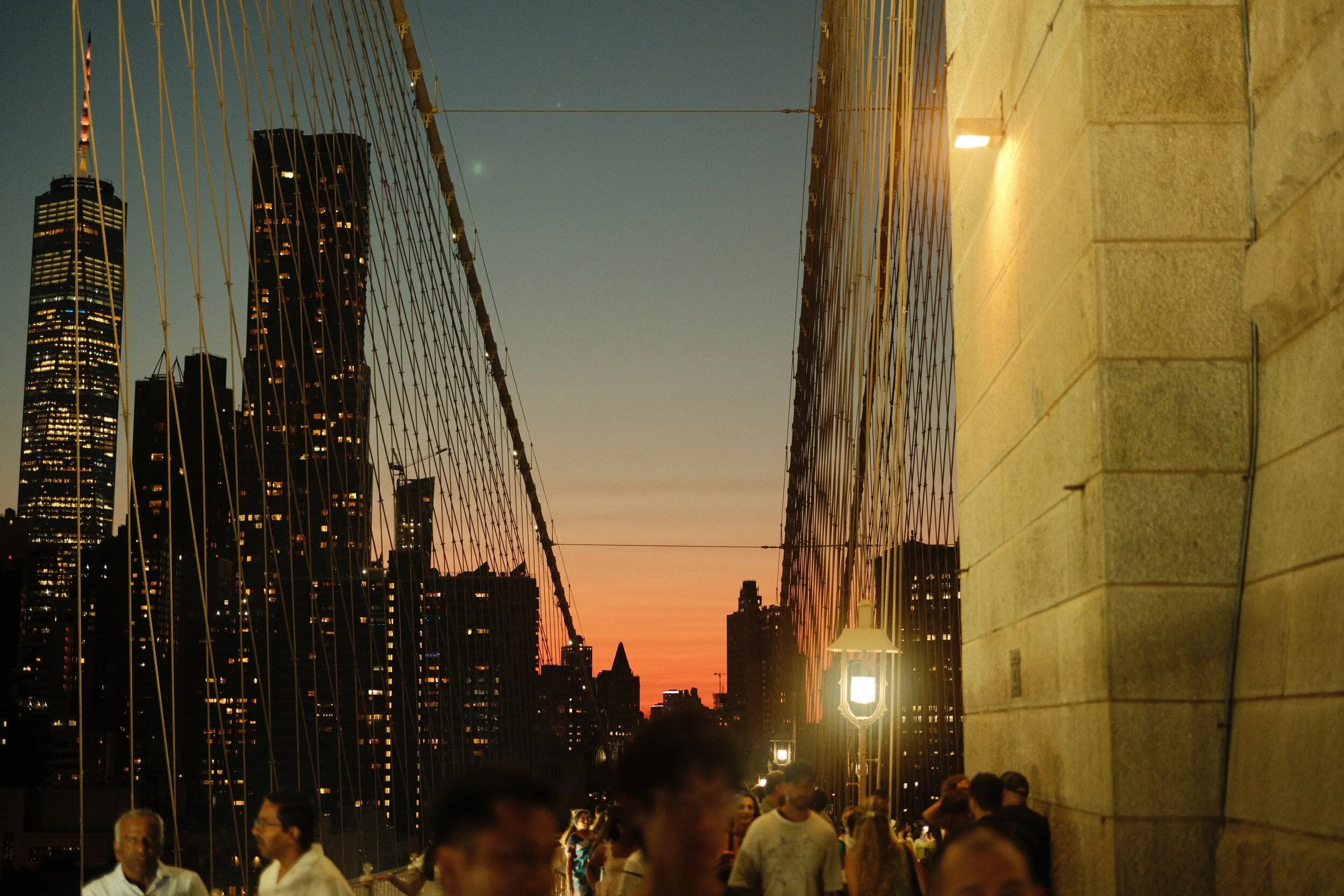 Nighttime scene on the Brooklyn Bridge with city skyline in the background and people walking along the bridge.