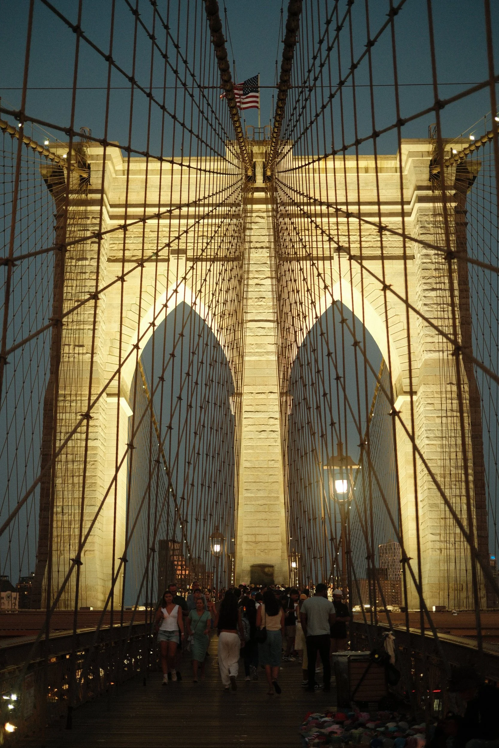View of the Brooklyn Bridge at dusk with an American flag on top and people walking on the bridge.