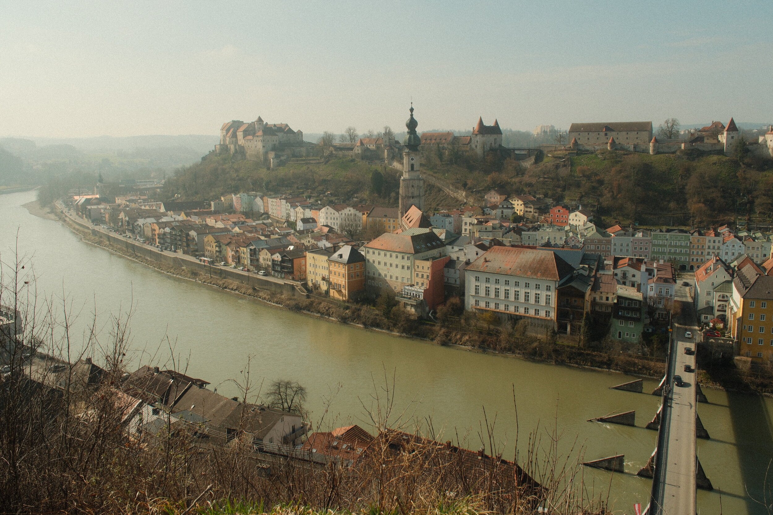 A scenic view of a European town along a winding river, with colorful buildings, a church with a tall spire, and a hilltop castle in the background overlooking the town.