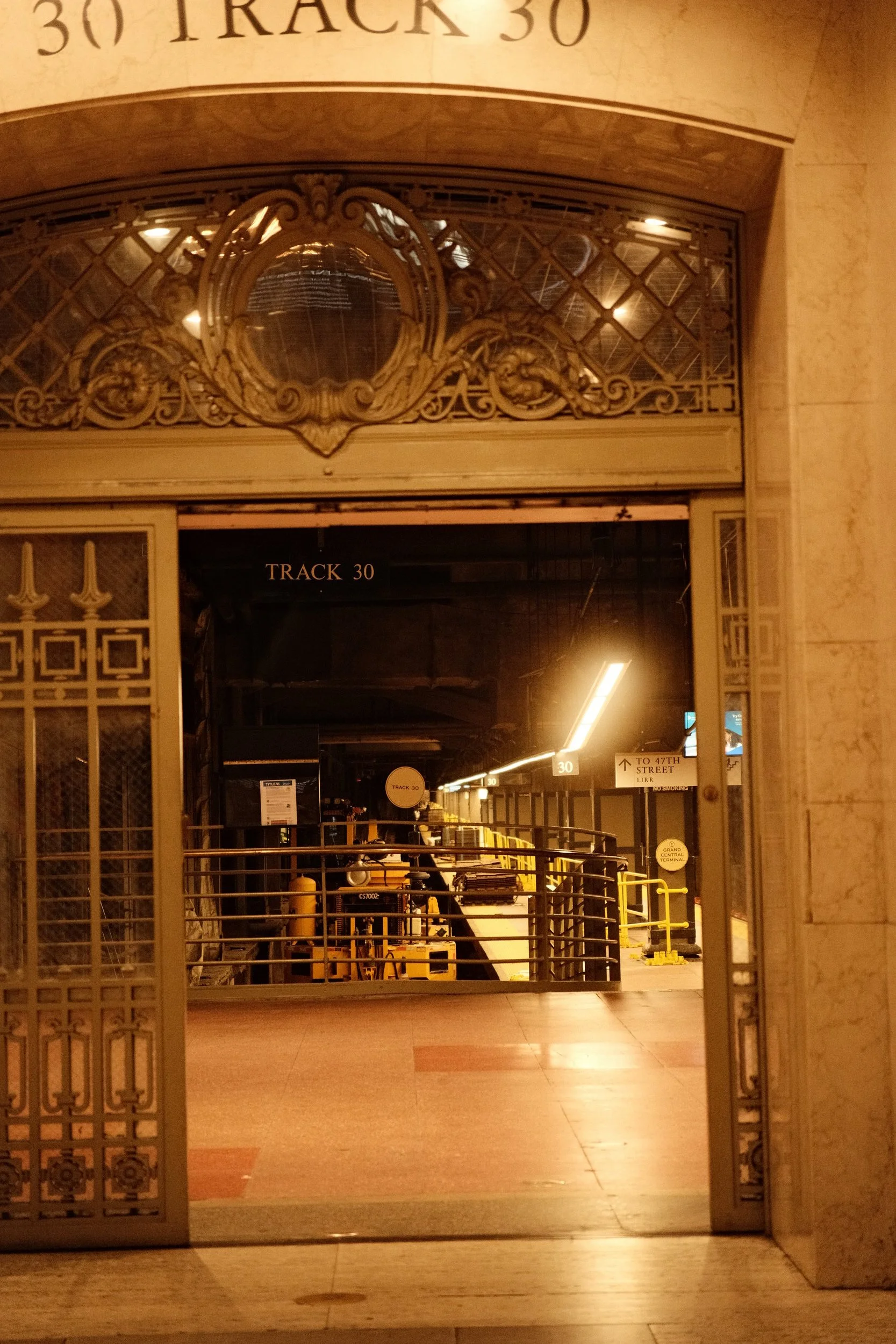 View of an entrance to a transportation area, specifically Track 30, with ornate metalwork and yellow carts or luggage trolleys inside. Signage indicates directions to 47th Street and Terminal.