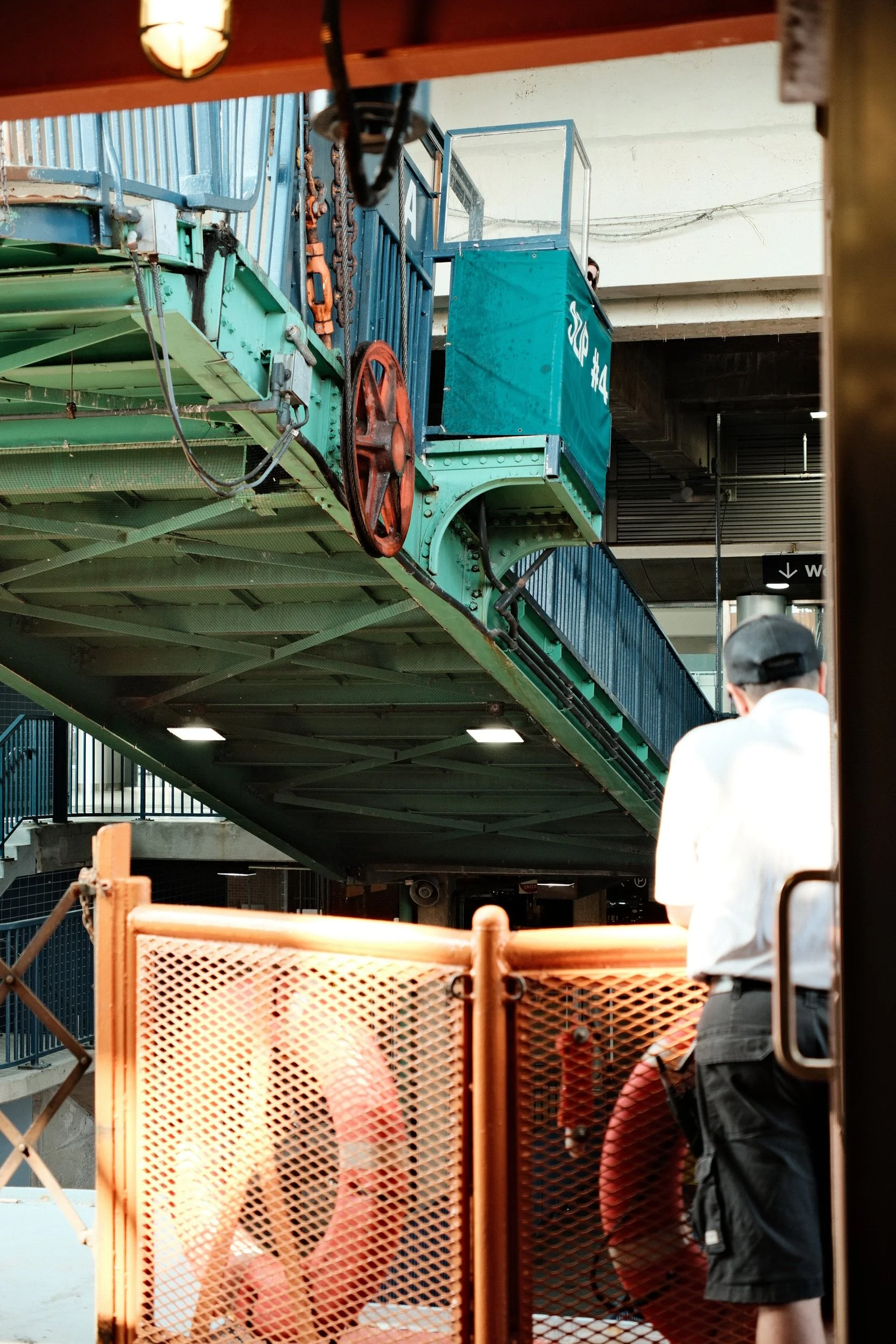 People walking on an elevated section of a city subway station. The station features teal and green painted metal structures and safety barriers in orange. A man in a white shirt and gray hat stands near the bottom right of the image.
