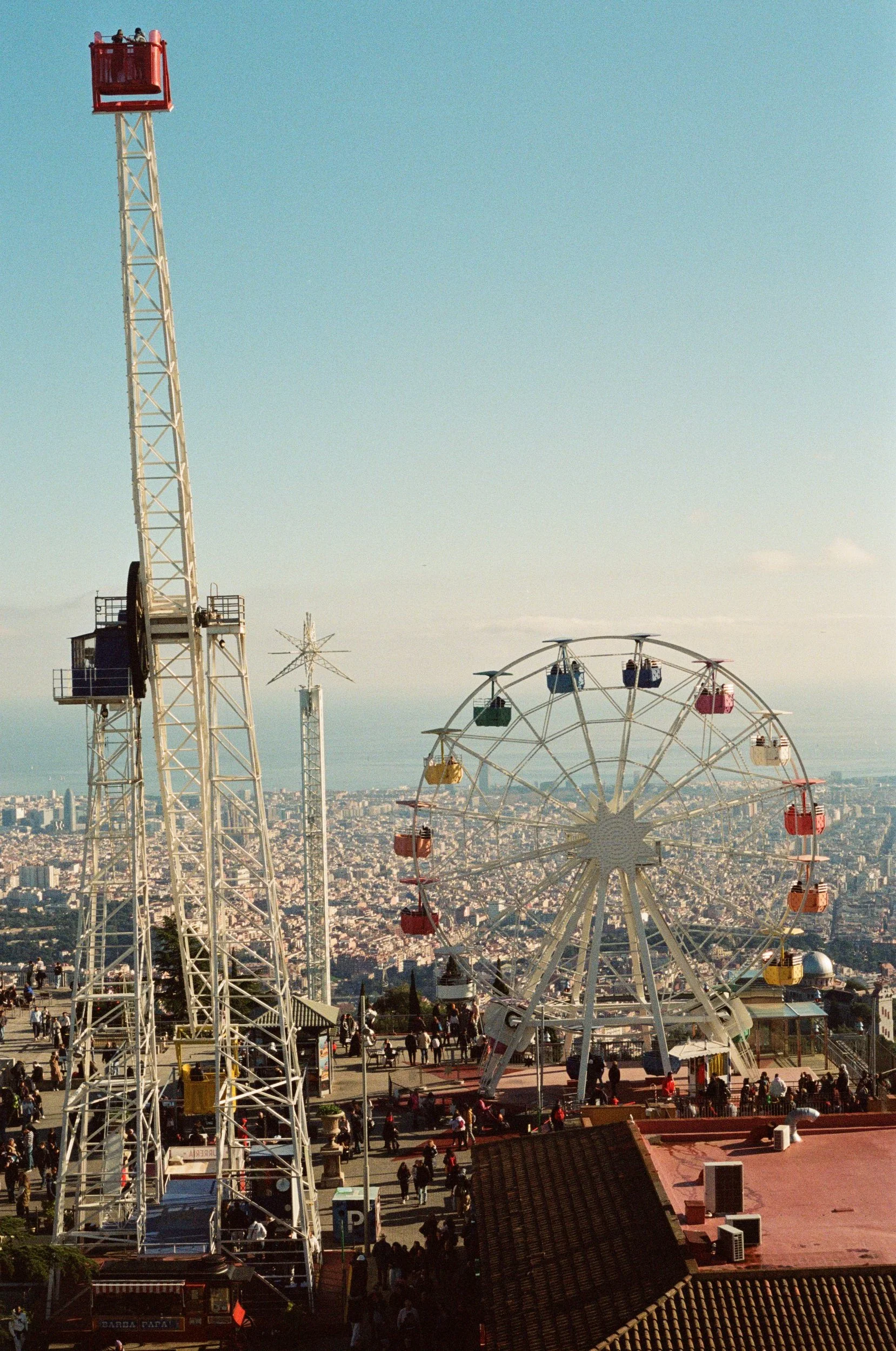 Amusement park with a Ferris wheel and a tower ride on a hill, overlooking a cityscape with many buildings and a body of water in the distance.