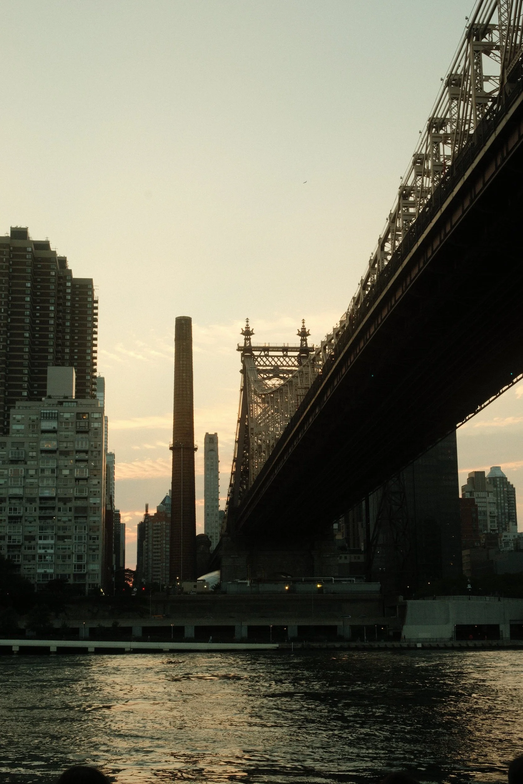 A city skyline at sunset with tall buildings, a bridge, and water in the foreground.