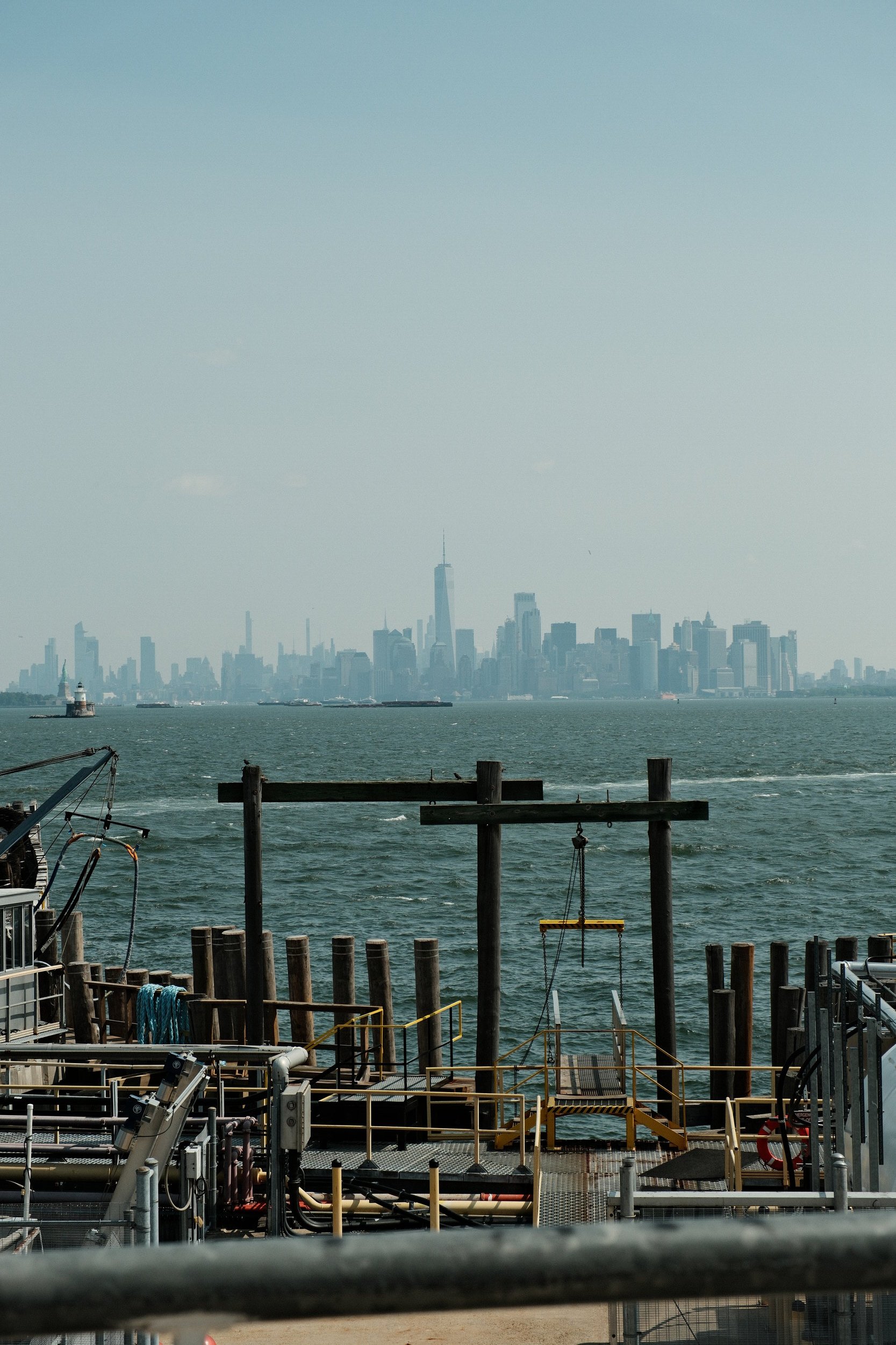 View of Manhattan skyline across the water, seen from a waterfront with industrial structures and wooden posts in the foreground.